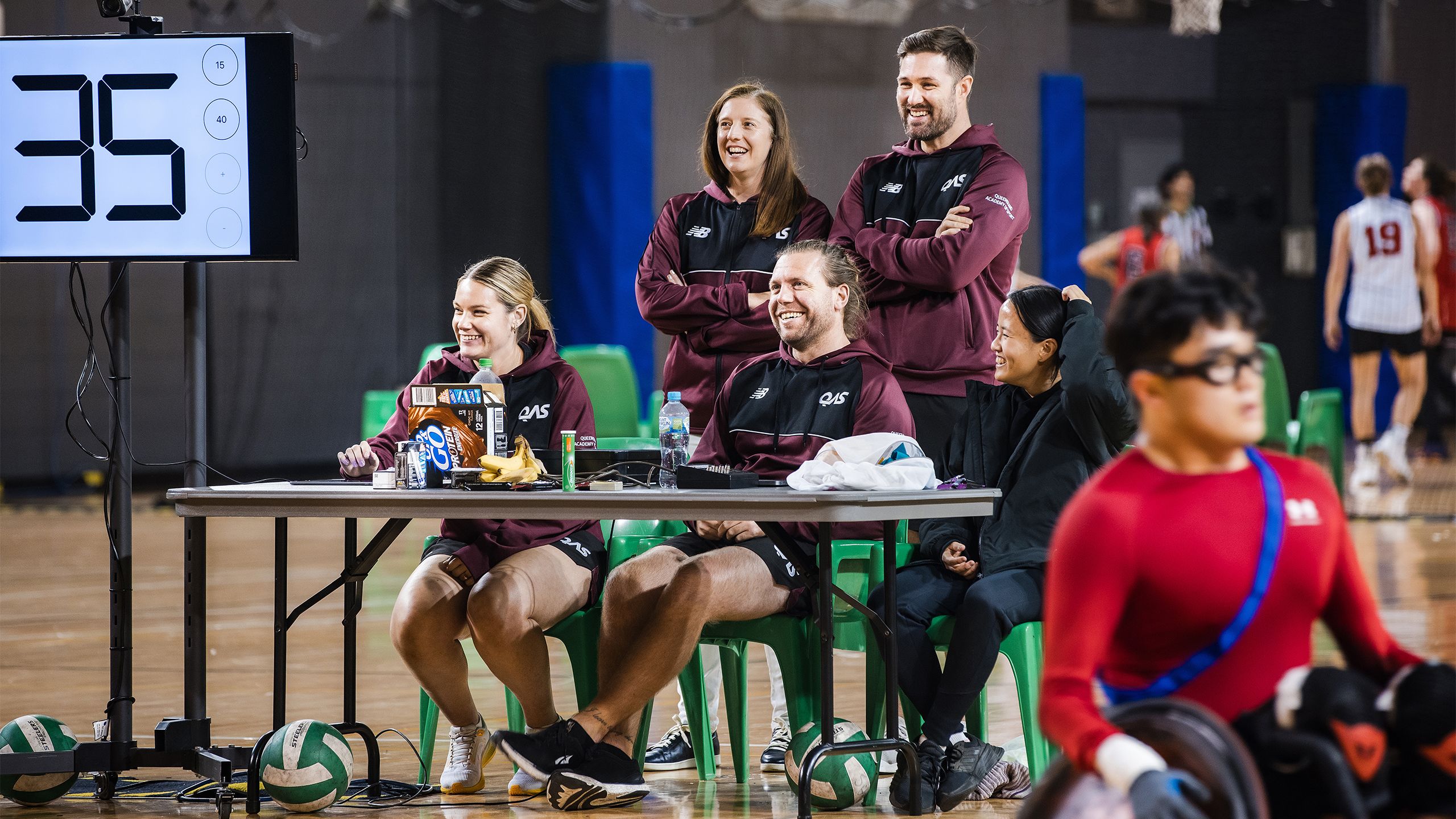 Five coaching staff sit at a table beside a hardwood court with a Para athlete in a wheelchair in the foreground.