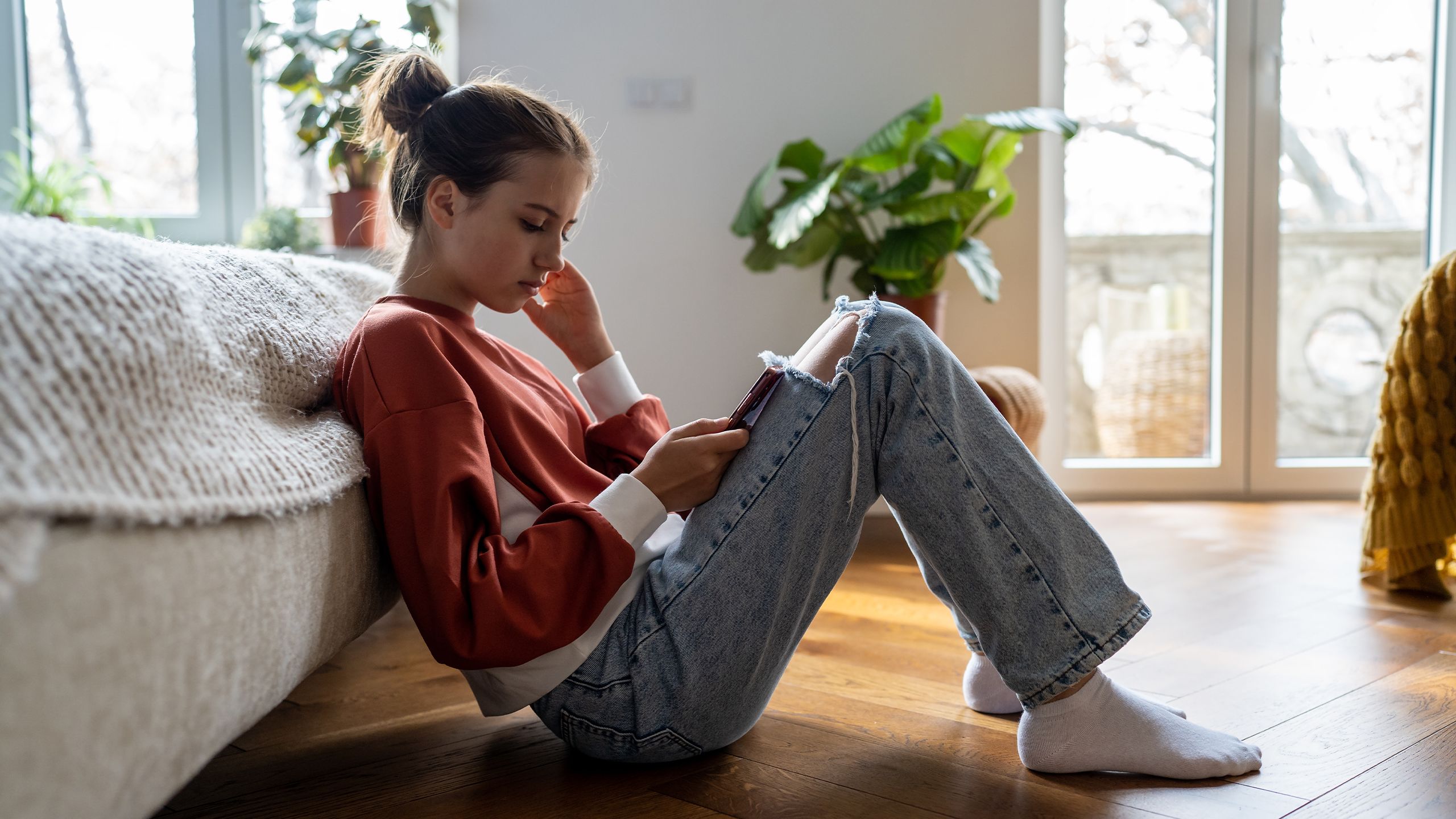 Teenage girl sitting on floor near bed using smartphone at home, scrolling social media.