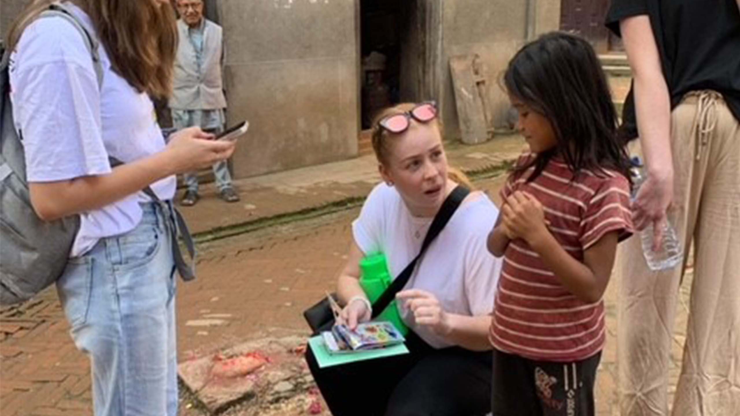 UQ student Pheobe Harrison talking with a young local child on streets of Nepal. The UQ student has red/blonde hair and is wearing a white t-shirt with sunglasses on top of her head. She is holding stickers as she talks to the child who is wearing a red shirt with white stripes.