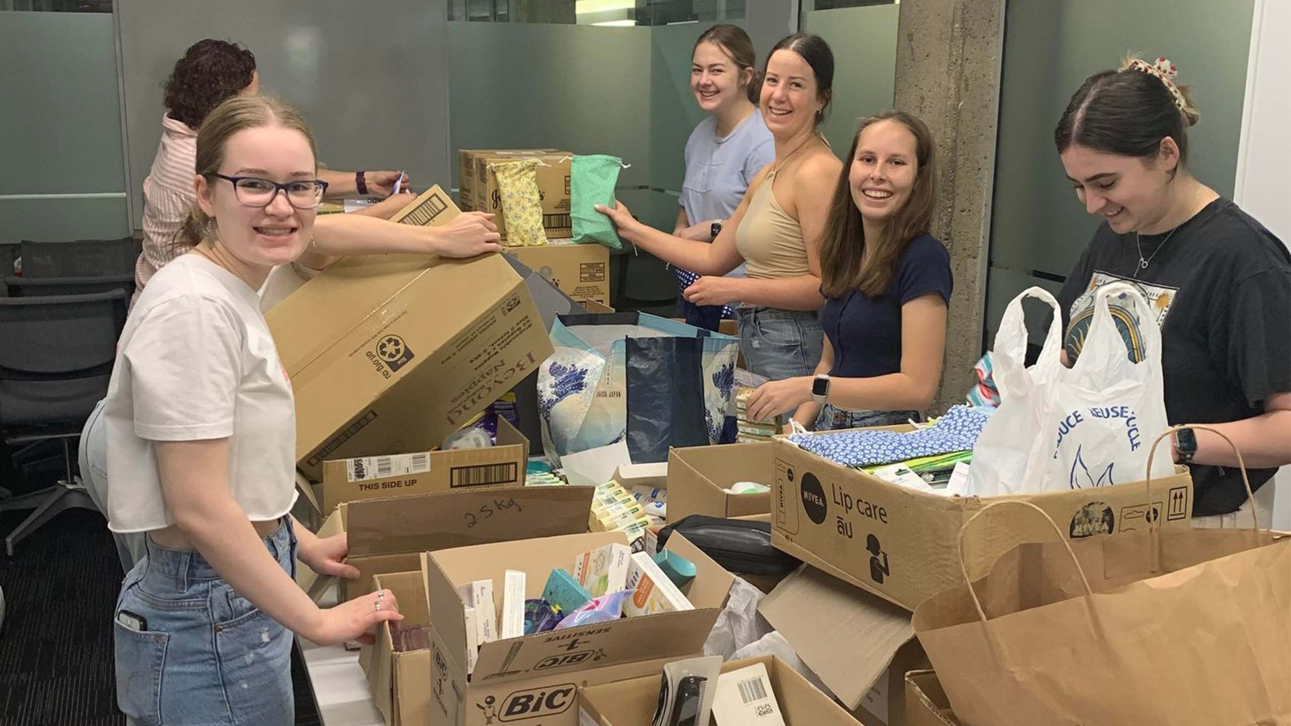 Five UQ students around a table with cardboard boxes filled with menstrual hygiene products. They are smiling at the camera as they pack