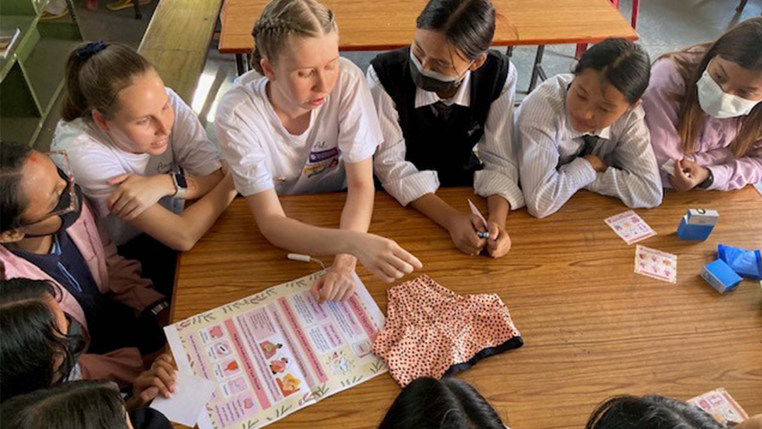 UQ students with Nepalese school girls sitting at a wooden table. There is a pair of women's underwear on the table next to a poster about menstrual health