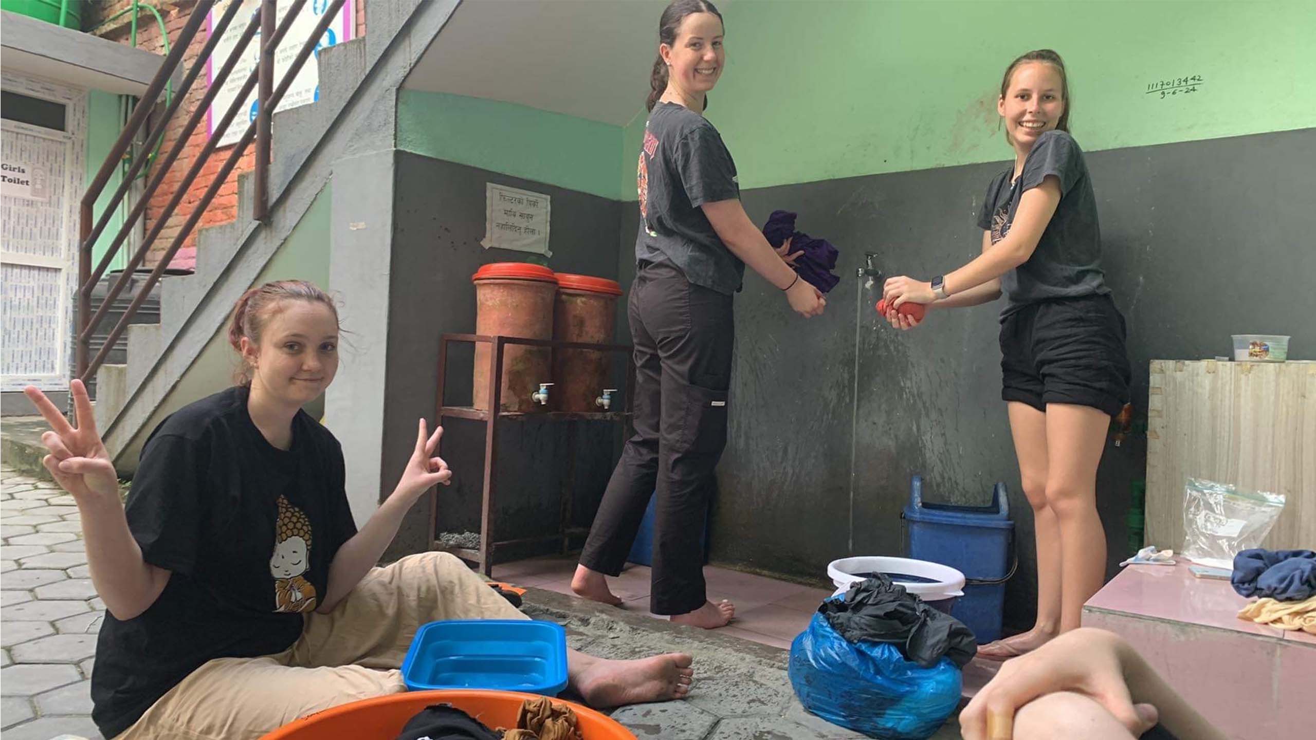 Three UQ students wash laundry using a single tap off the wall and tubs. Two are rinsing their clothes while smiling at the camera, the other is sitting on the floor doing the 'peace sign' with her hands while smiling.