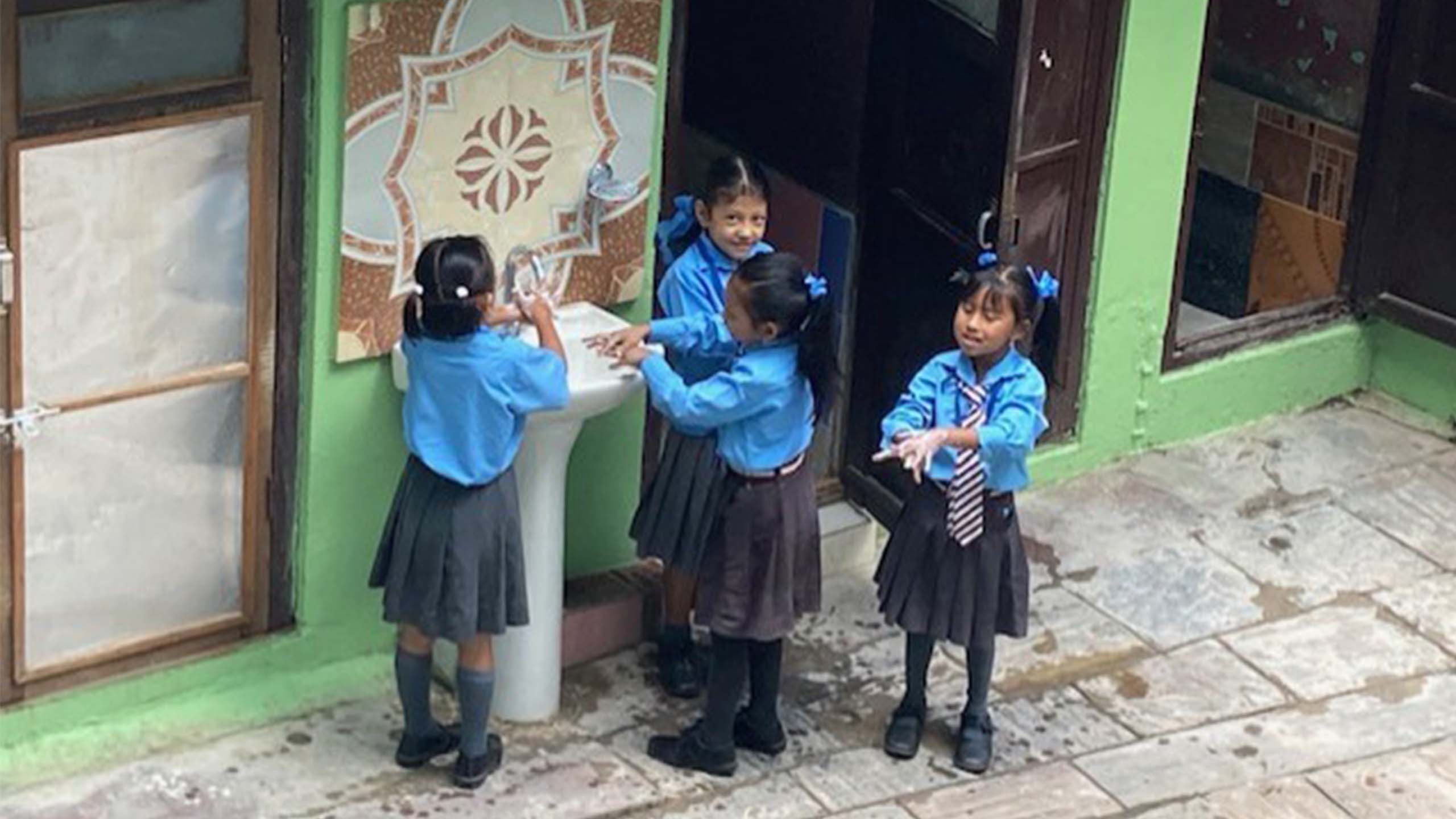 Four young Nepalese girls washing their hands at an outdoor sink at their school. they are wearing uniforms with blue long sleeve shirts, grey pleated skirts, long grey socks with black shoes and blue ribbons in their hair.
