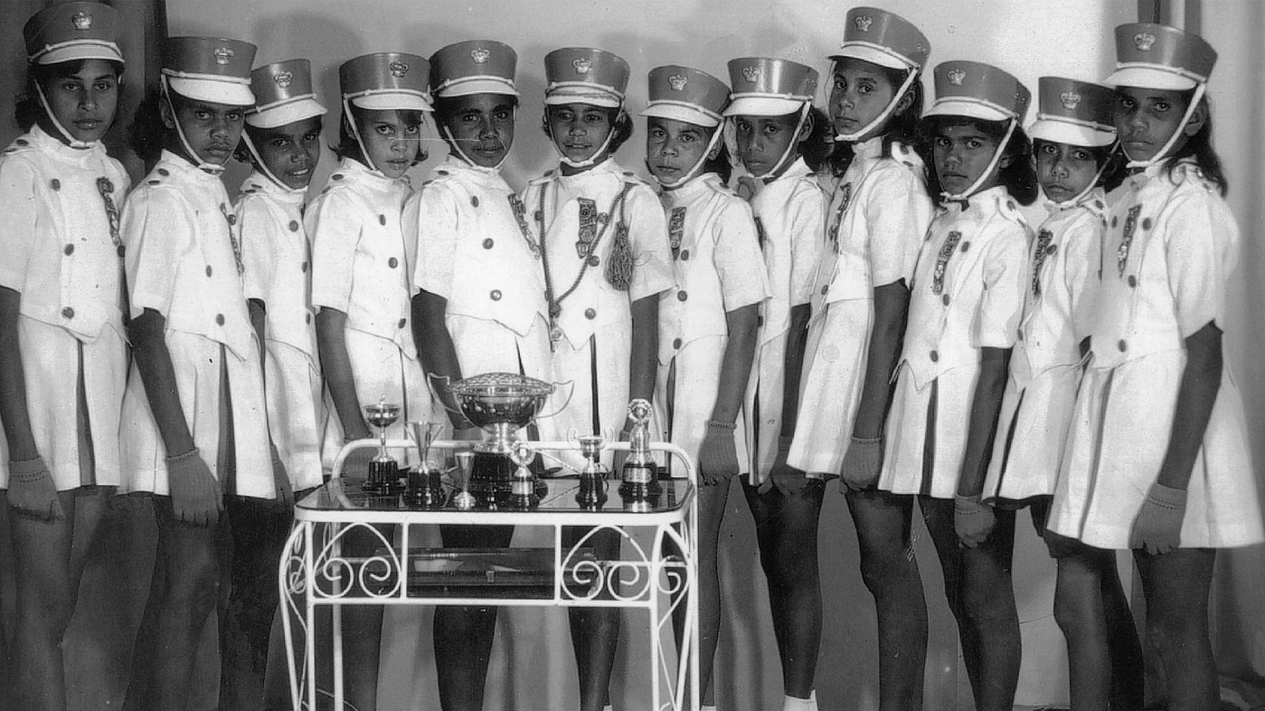 A marching band made up of young Aboriginal Australian girls standing behind a trophy. The image is in black and white.