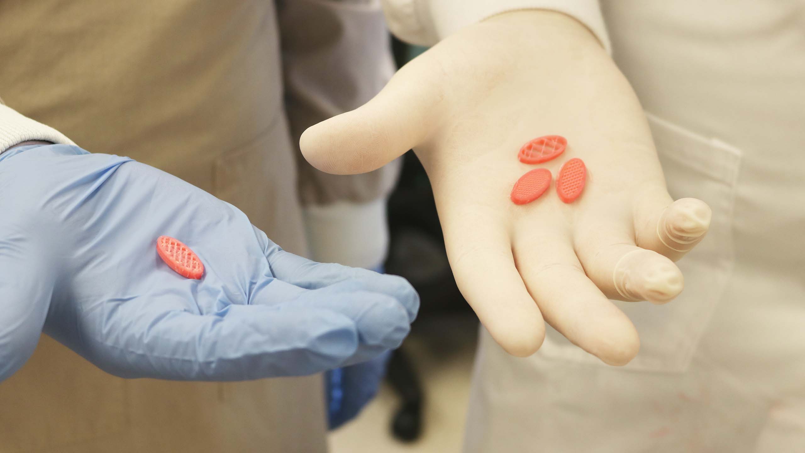 A close up of two gloved hands holding 3D-printed pills. The pills are orange and an oval shape.