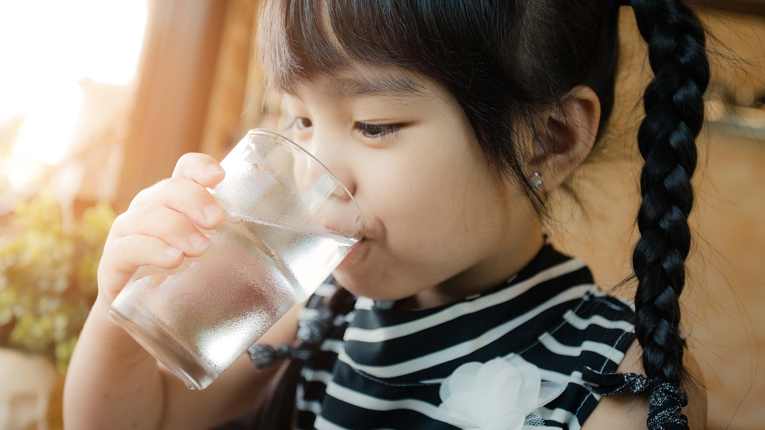 A young girl drinking a glass of water. She has black hair in two braided ponytails.