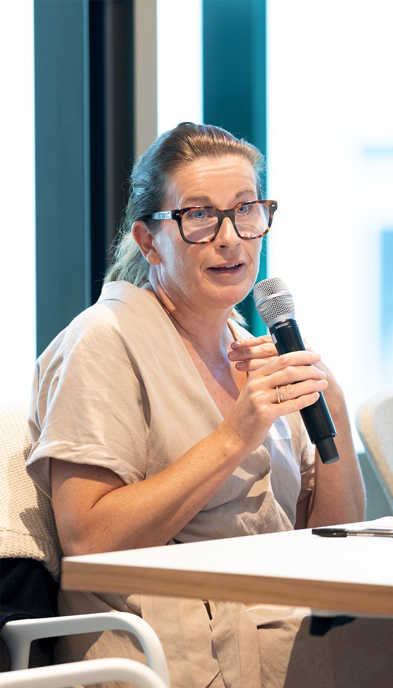 Woman sitting at a table speaking on a microphone. She has light brown hair, black glasses and in wearing a light tan outfit.