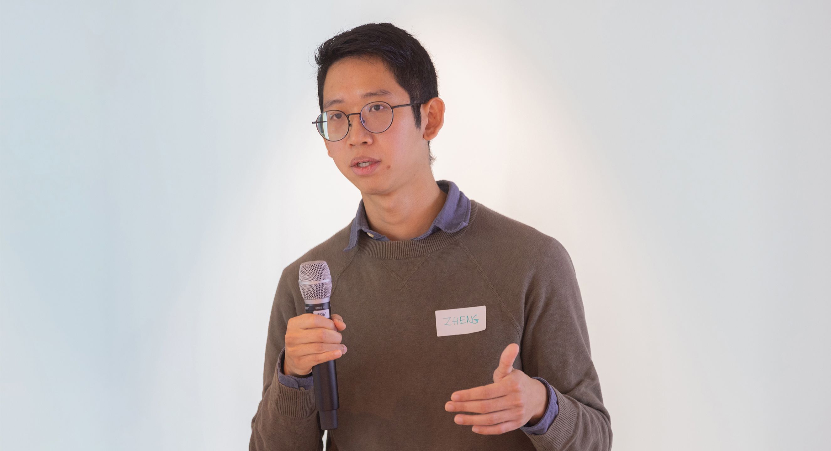 Man speaking on microphone. He has dark hair, thin rimmed glasses and is wearing a brown sweater with a blue collared shirt underneath