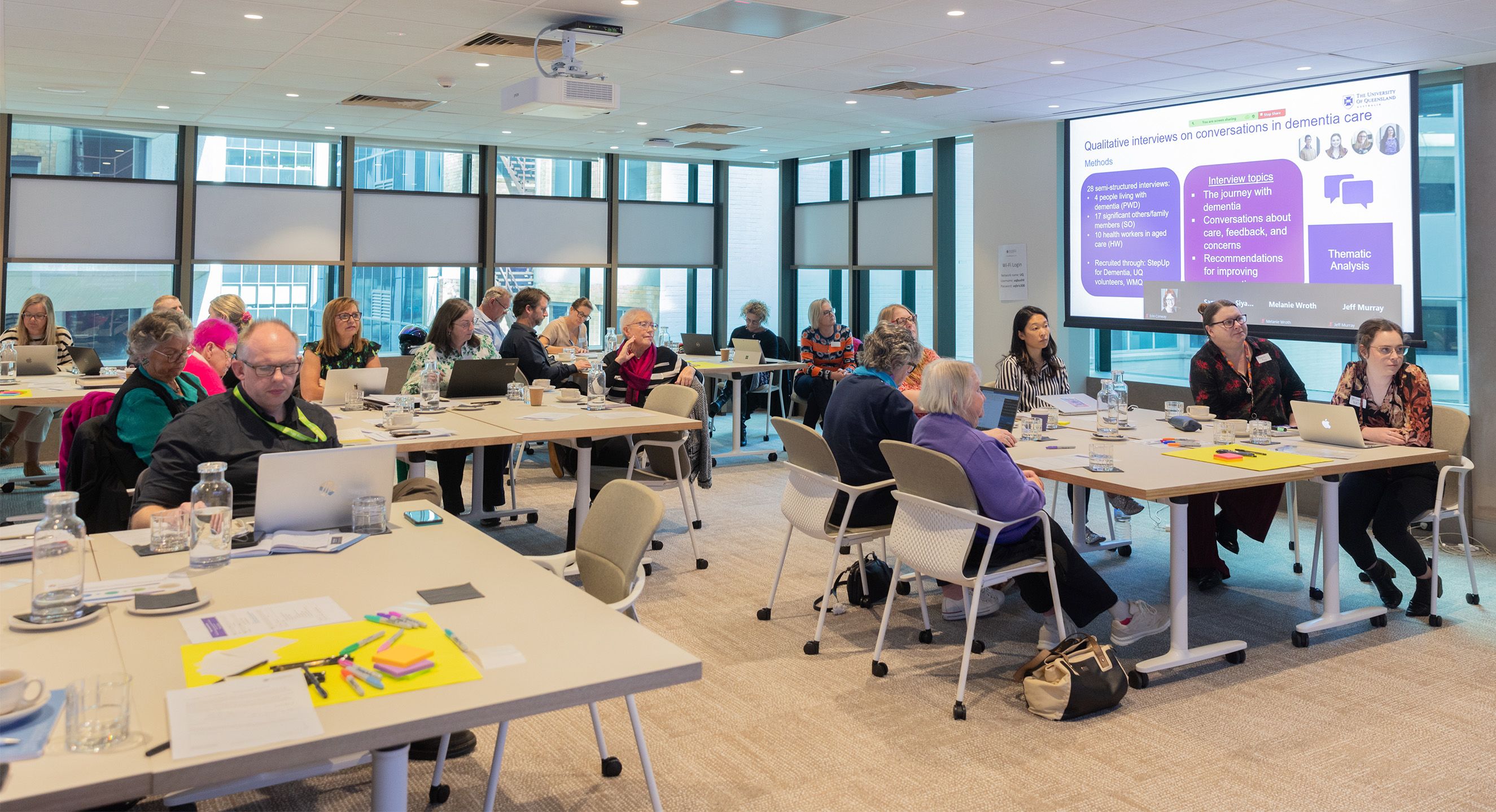 People sitting in groups at tables listening to a presentation. There are stationary items on the tables and a powerpoint presentation on a hanging screen.