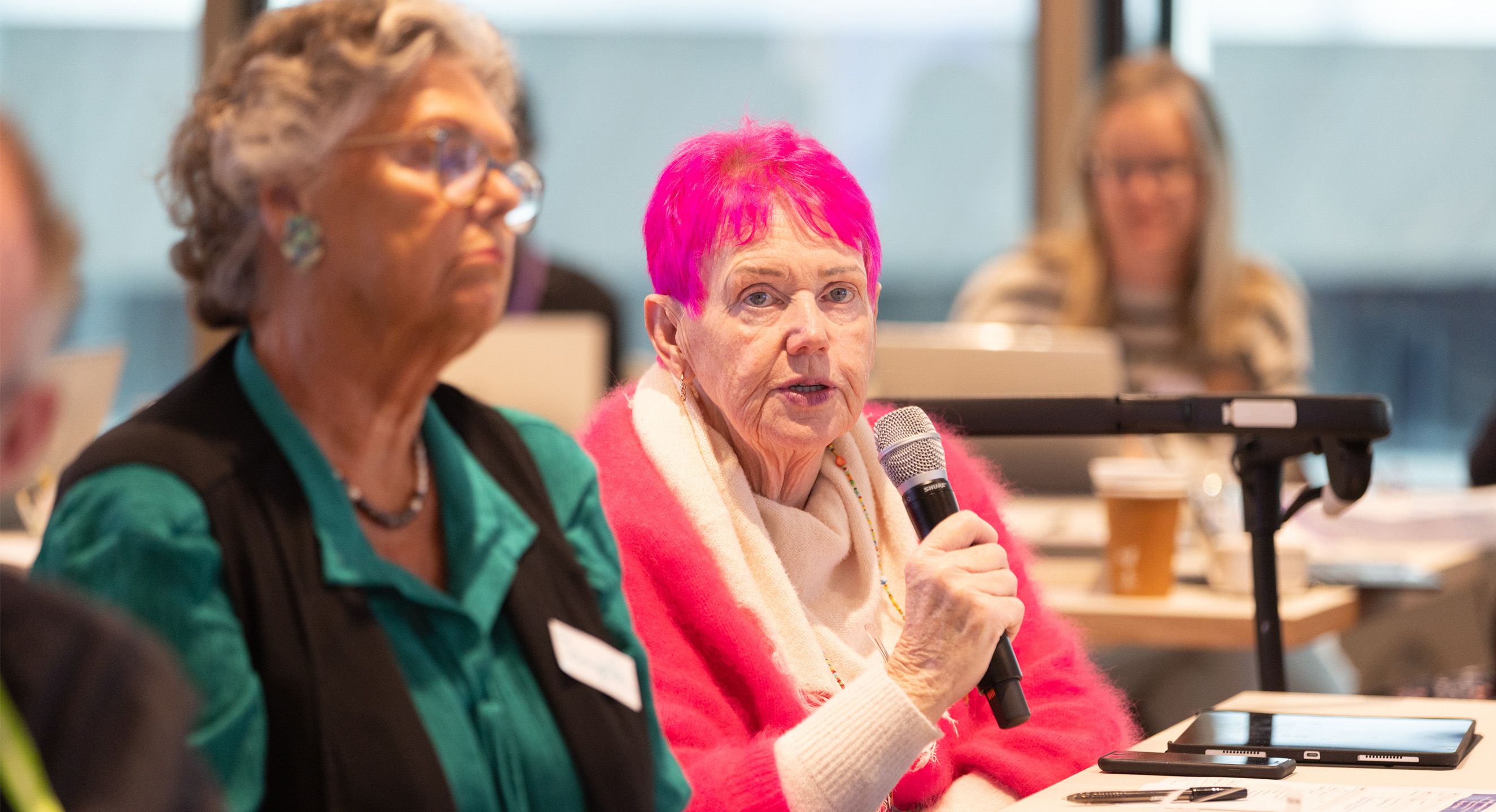 Older woman with vibrant pink hair speaking on microphone. She is wearing a fluffy pink sweater with a beige scarf