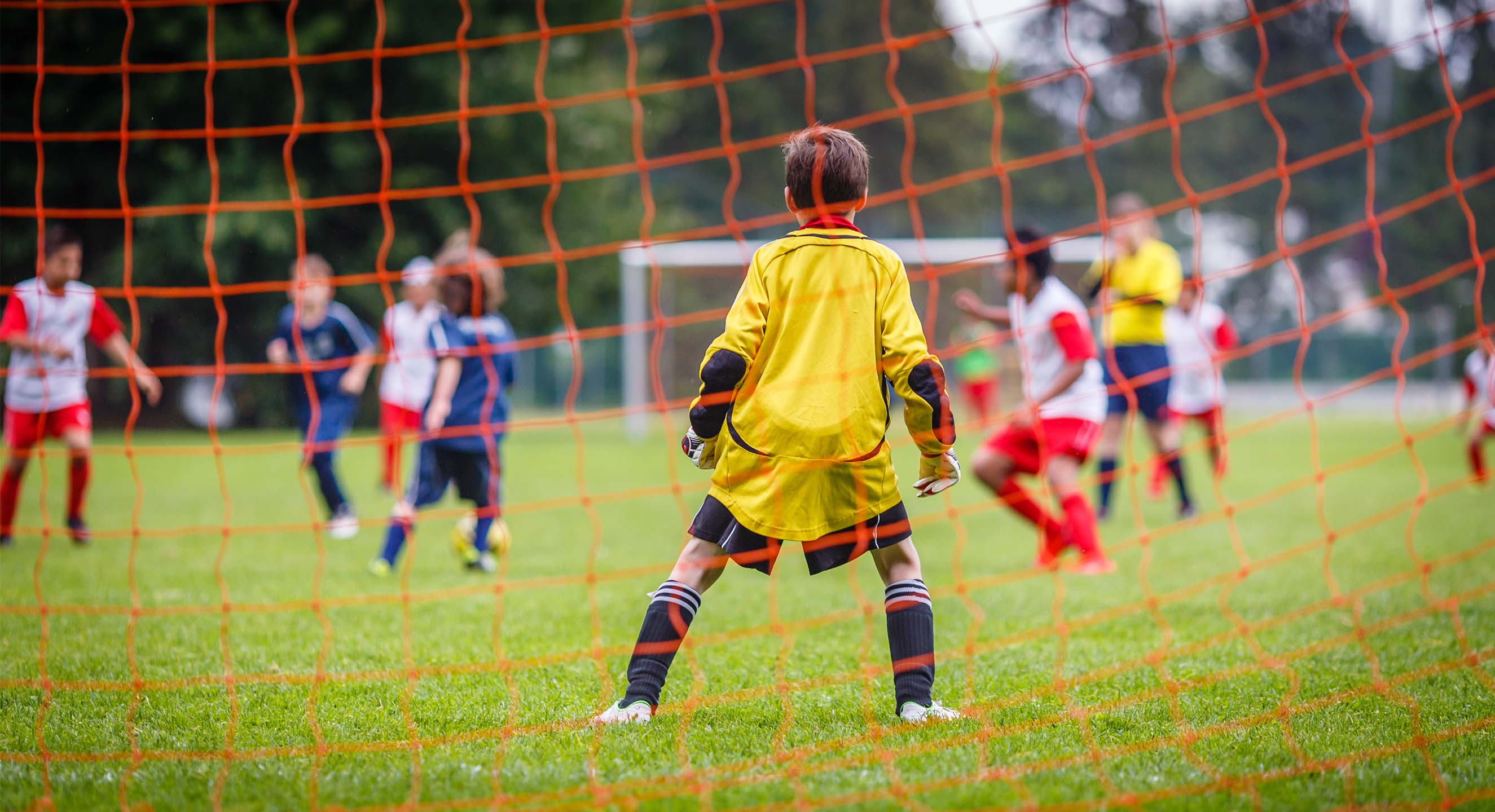 A young boy standing in a soccer goal. He is watching the other players on the field approaching the goal