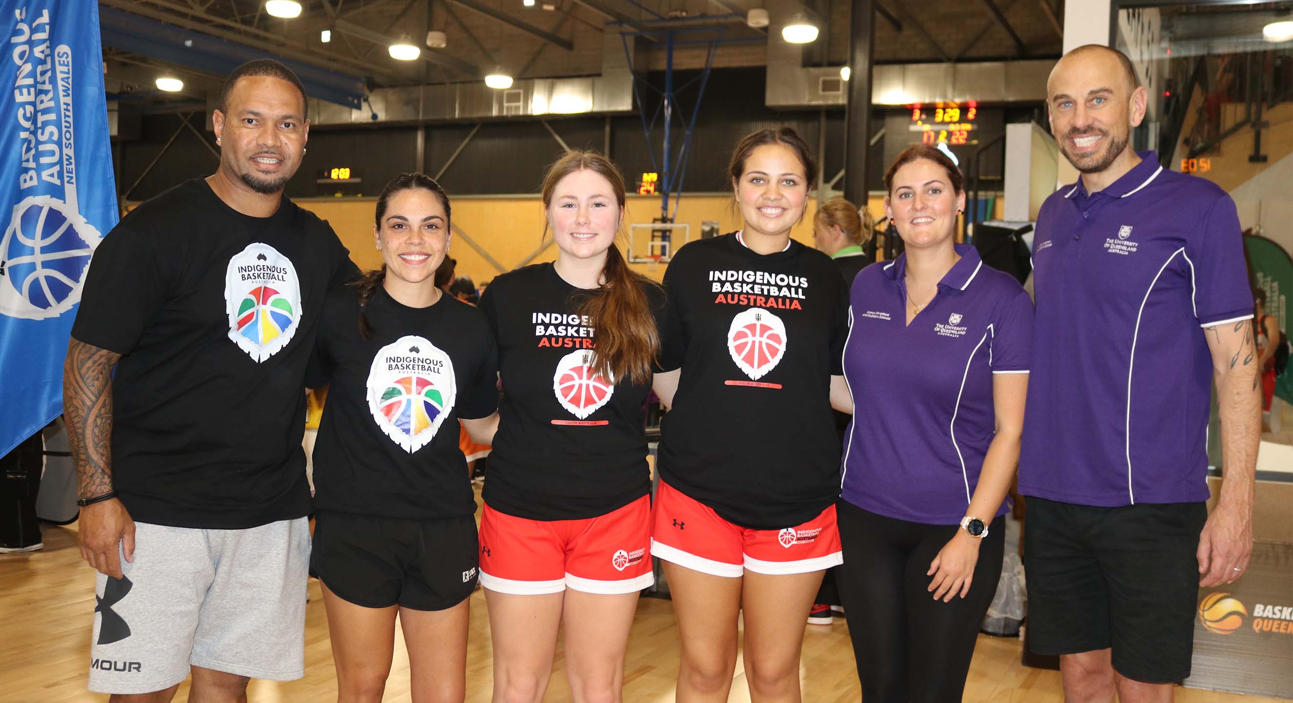 Six people, two wearing purple polo shirts and two wearing Indigenous Basketball Australia uniforms, standing on a basketball court in a line and smiling