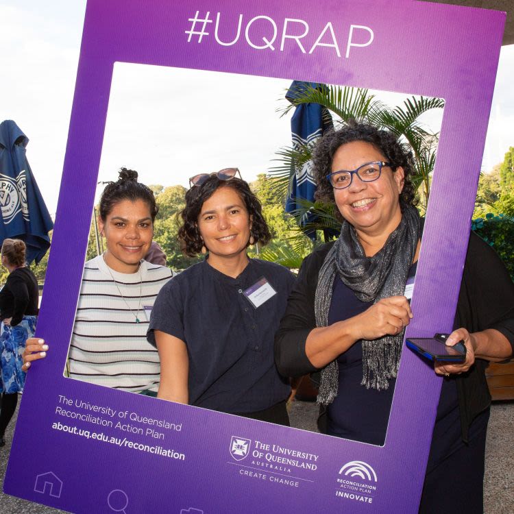 Three women smile and hold up a purple Instagram frame that says #UQRAP.
