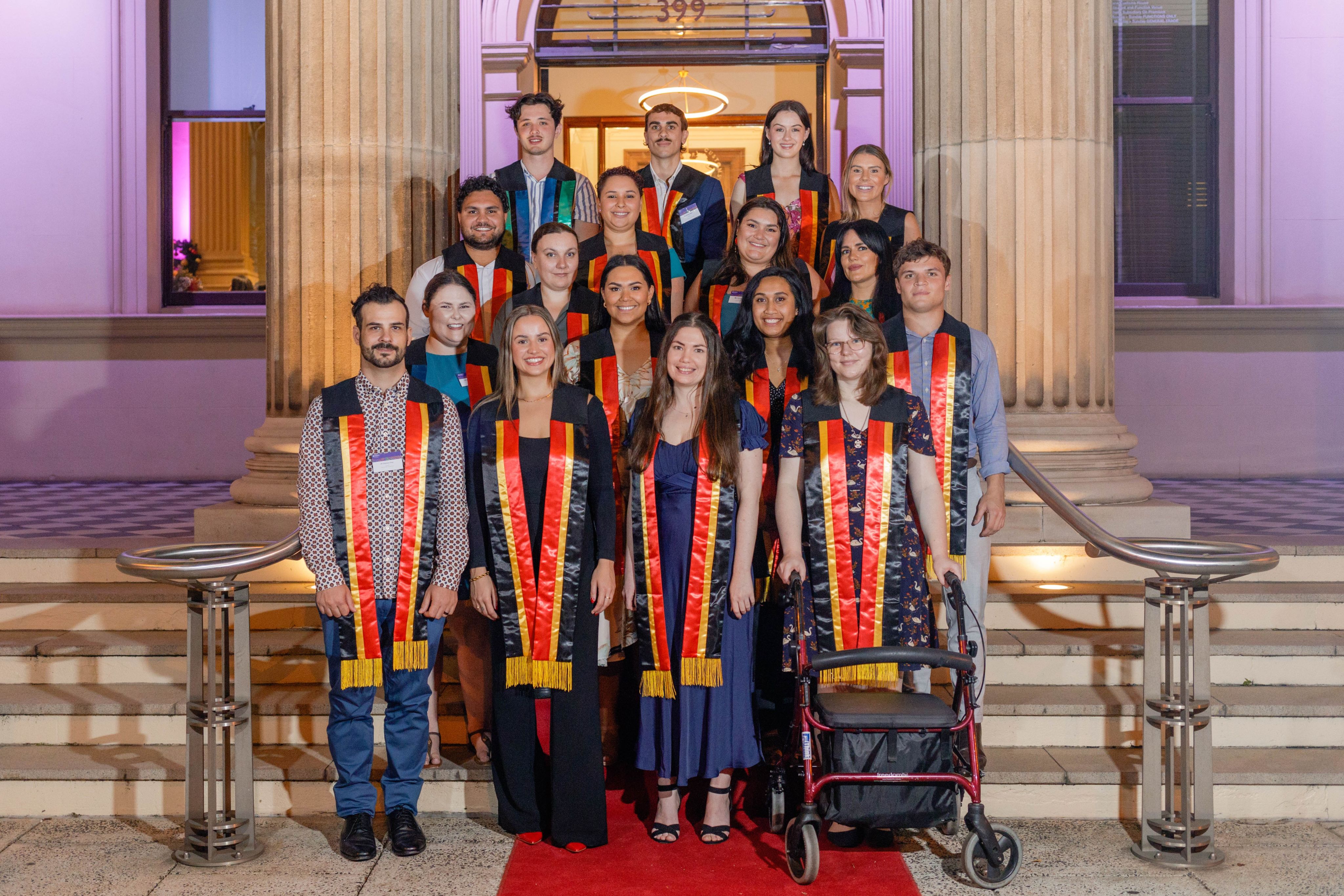 A group of 17 Aboriginal and Torres Strait Islander students wearing sashes over their shoulders while standing in rows on the steps of an old sandstone building