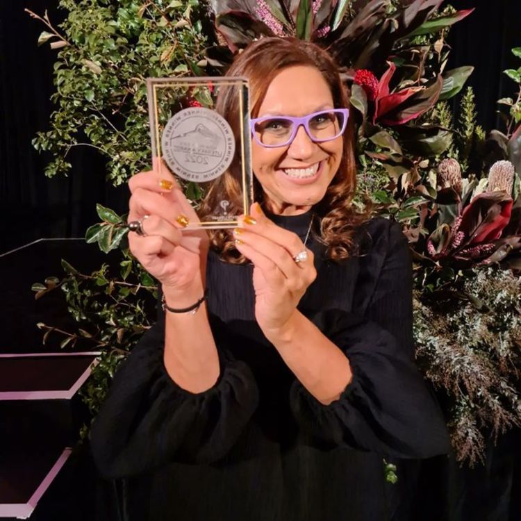 Dr Anita Heiss, a woman with light brown hair wearing lilac glasses, holding a rectangular glass trophy and smiling