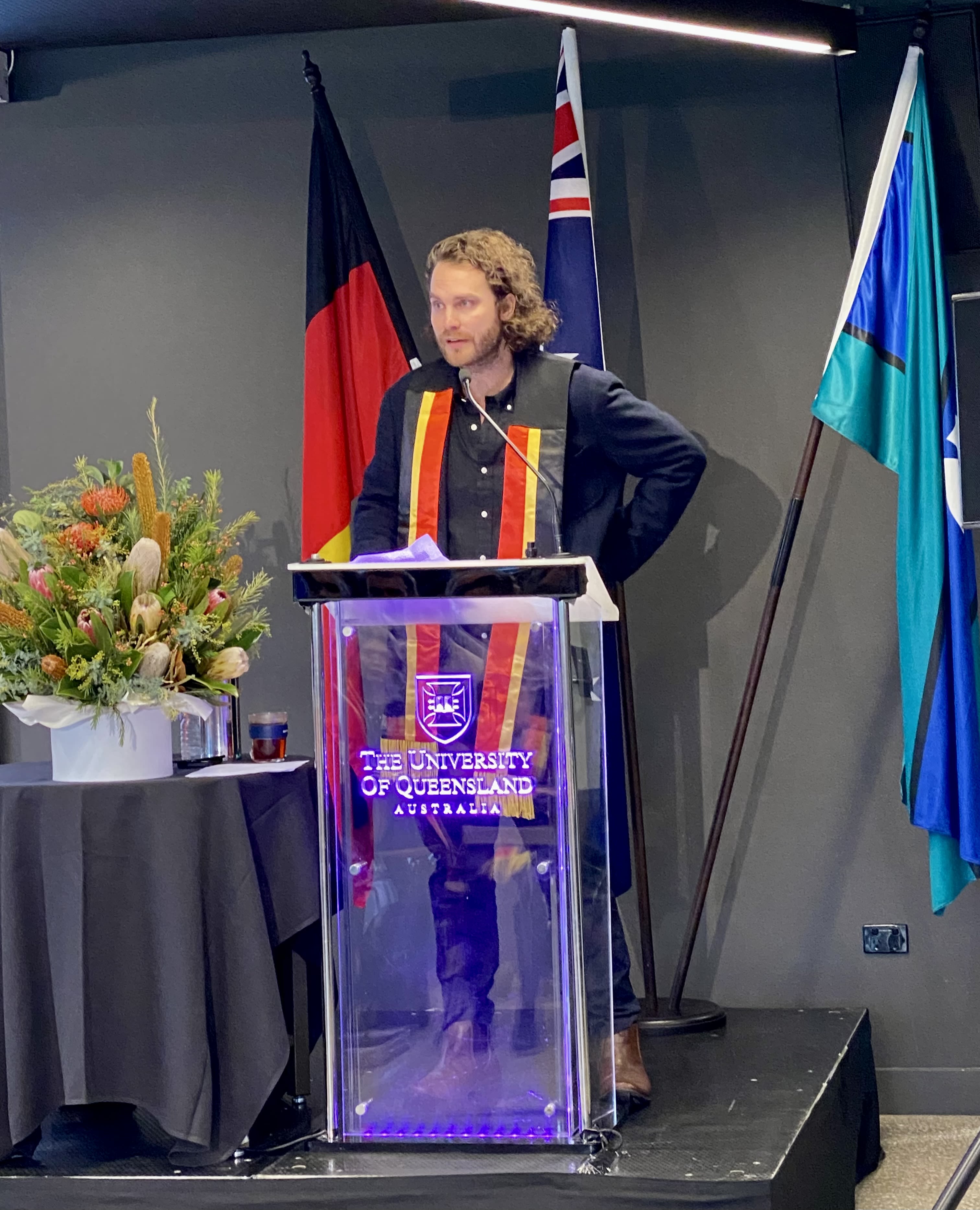 Mitch Rom, a young man wearing an Aboriginal Sash, speaking at a lectern with a microphone