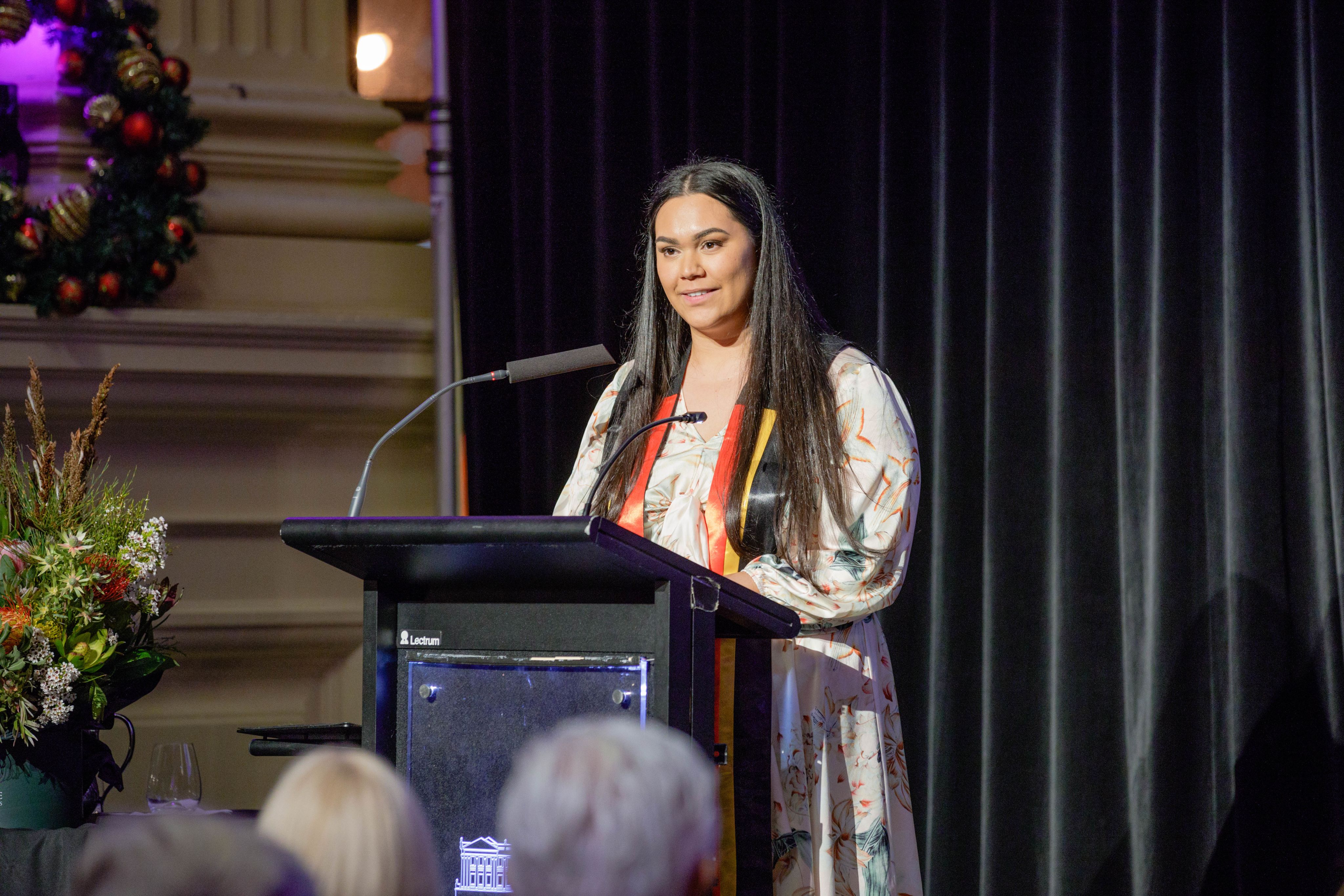 Dr Ella Ceolin speaking at a lectern while wearing an Aboriginal sash over her shoulders
