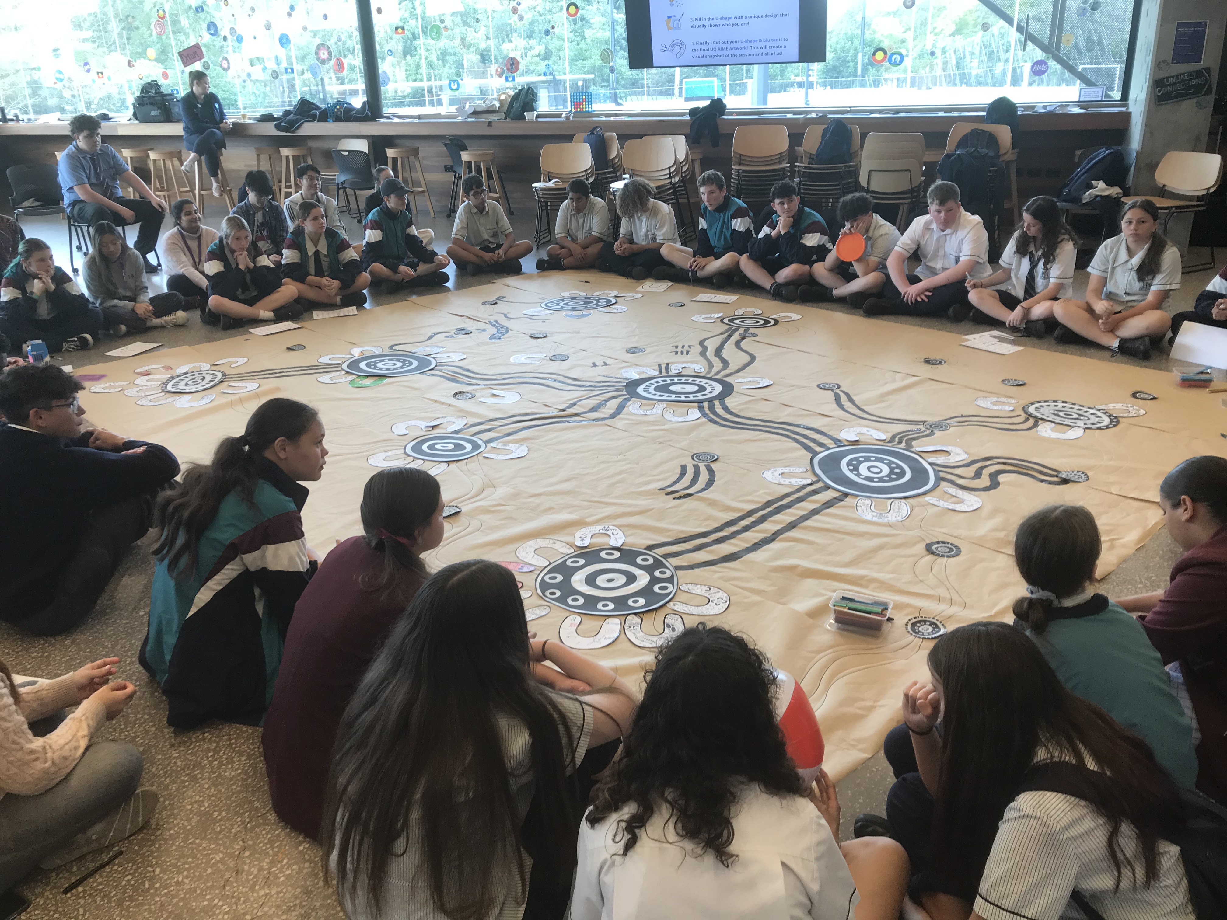 A group of 40 school students wearing their uniformssitting on the ground surrounding a large rectangular piece of artwork