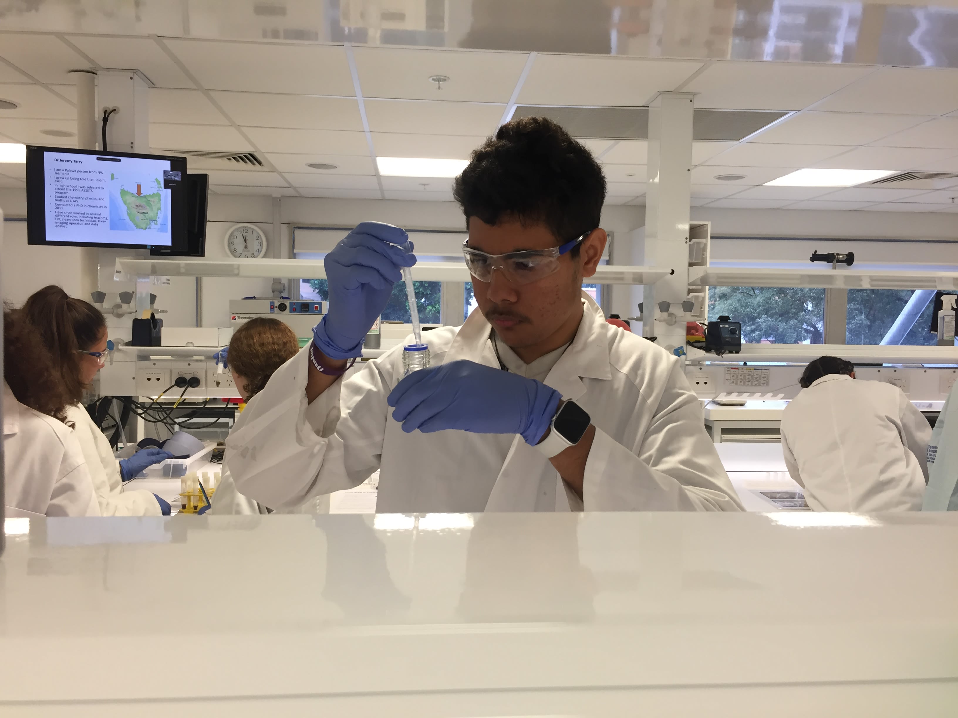 A teenager wearing protective equipment using a pipette to transfer liquid into a small container in a science laboratory