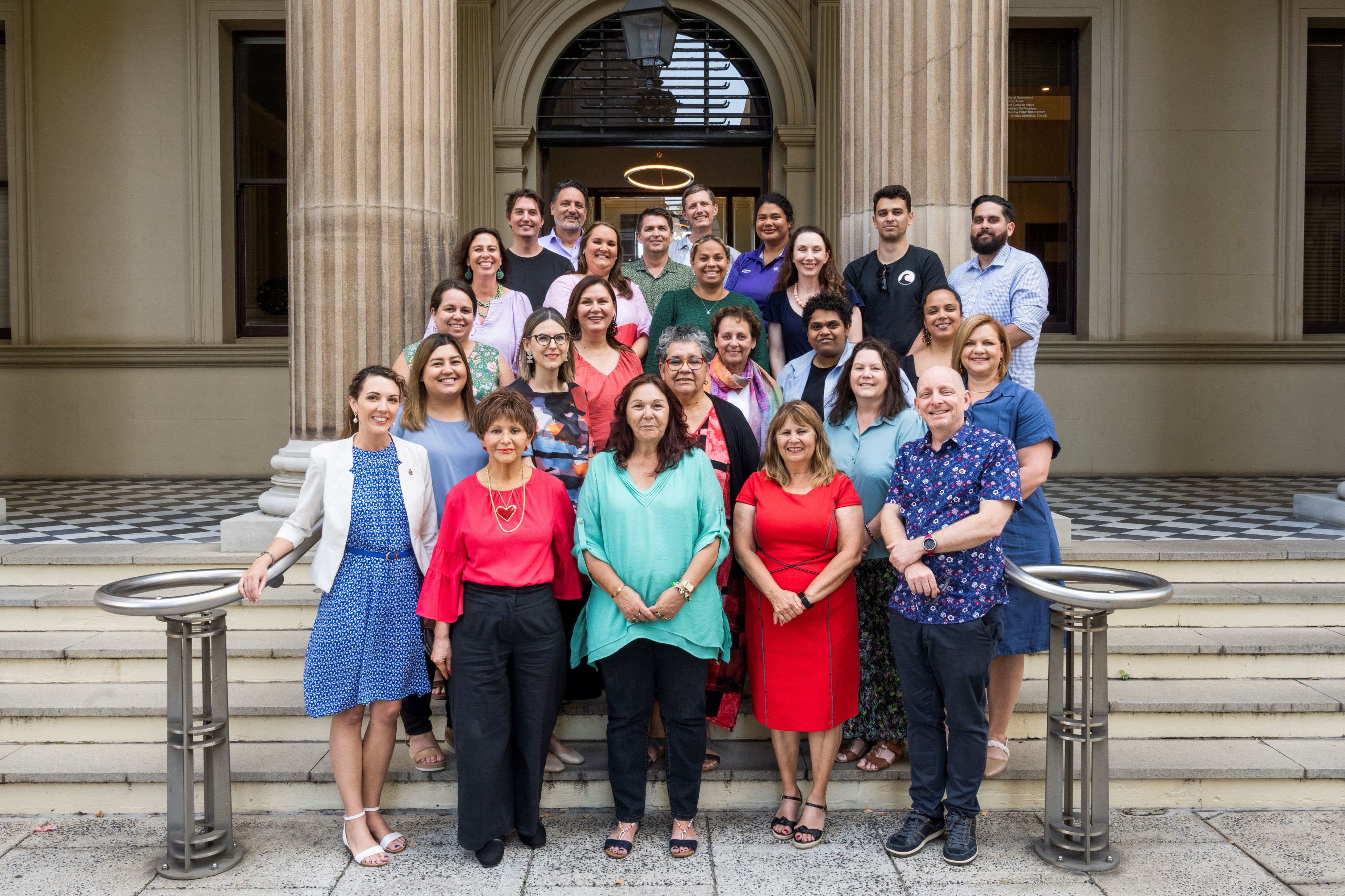 A team of 20 people standing in rows on the front steps of a sandstone building
