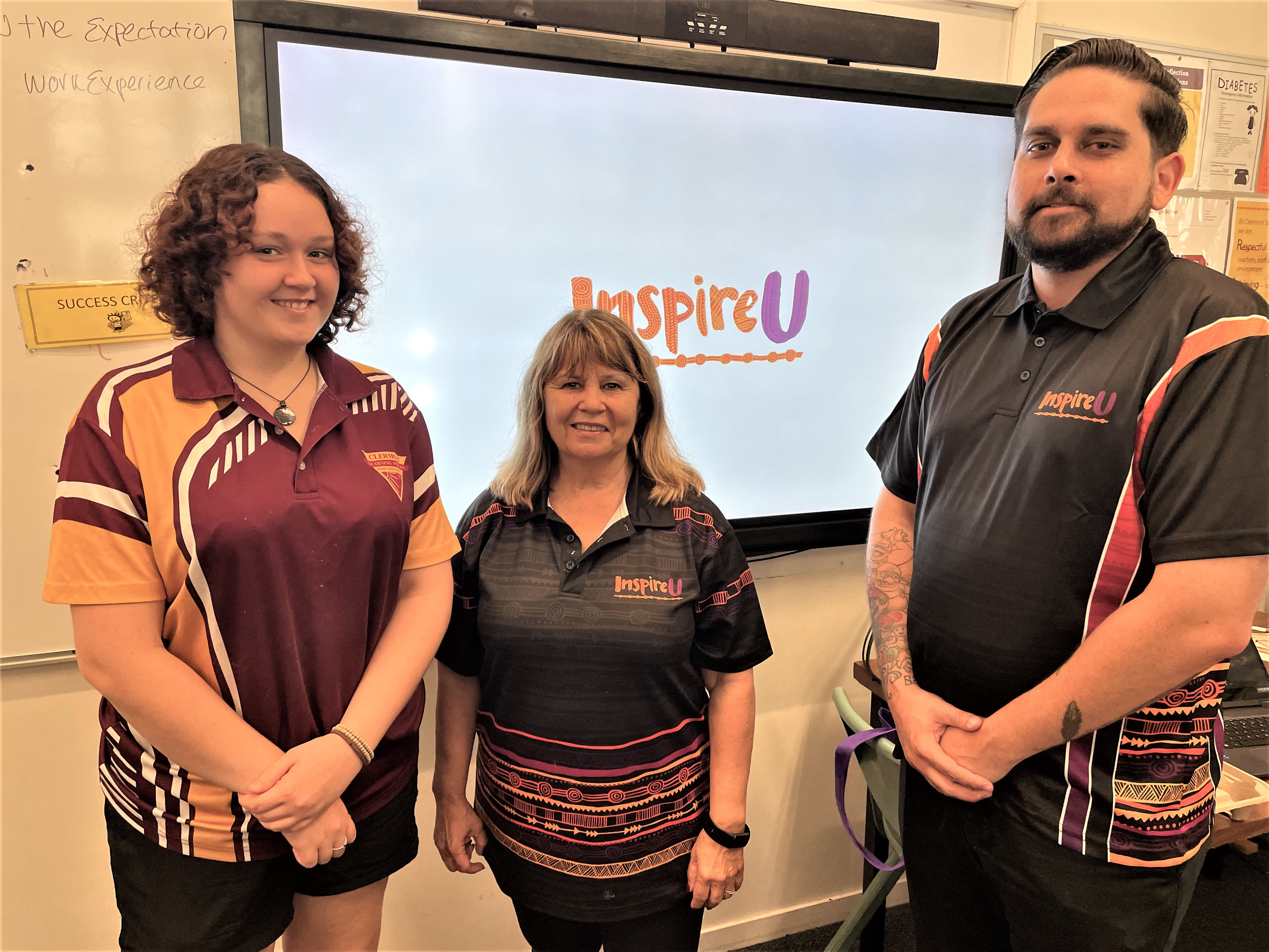 A school student in uniform and two adults wearing InspireU shirts standing in front of a large screen in a classroom