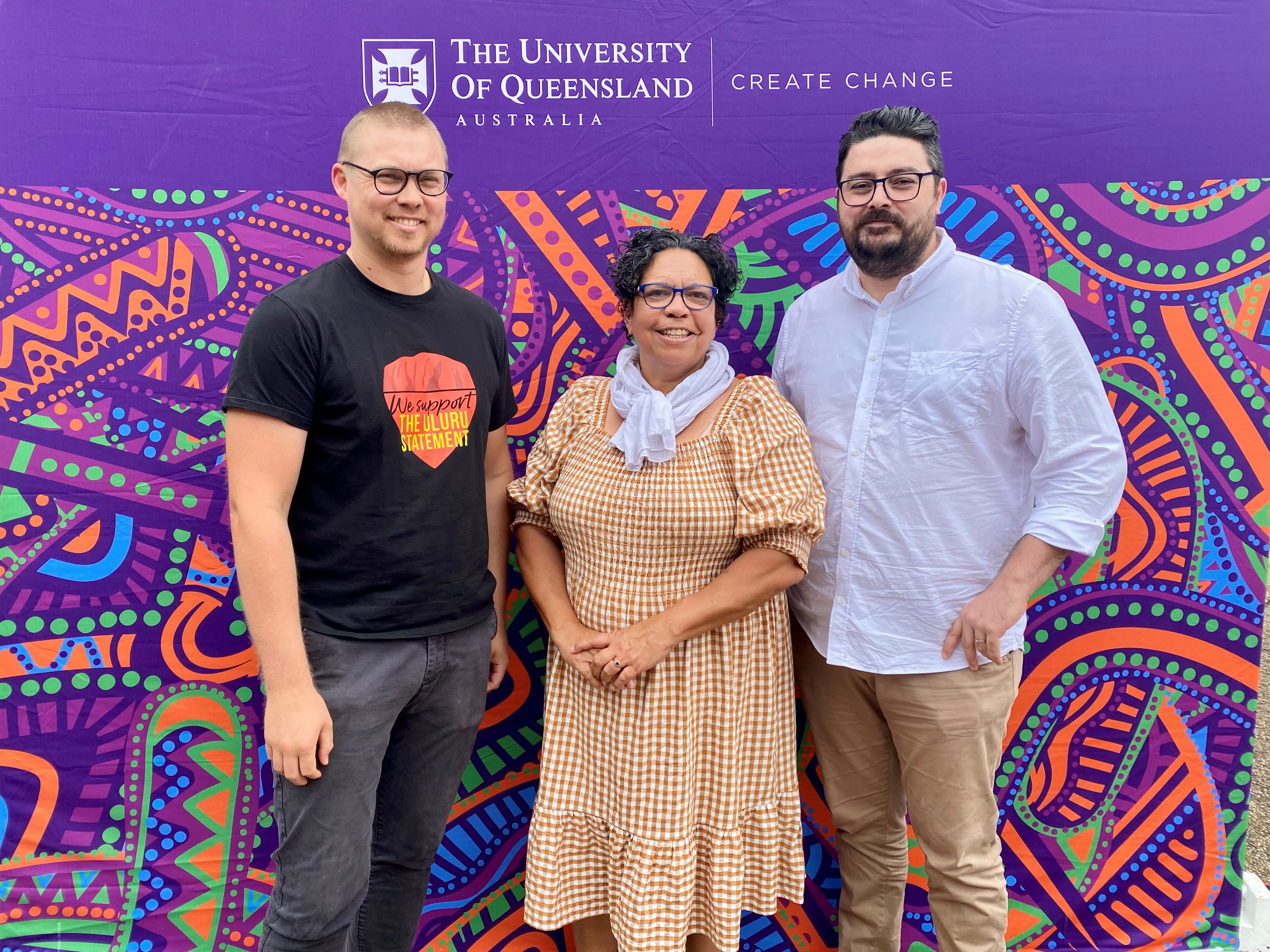 Three people standing in front of a sign with the University of Queensland logo and colourful Indigenous artwork inspired by the Brisbane River