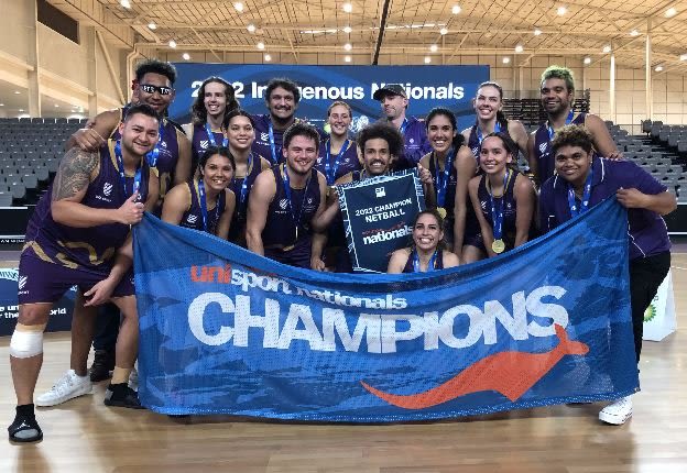 A mixed-gender netball team wearing uniforms and holding a blue banner that says 'UniSport Nationals Champions'