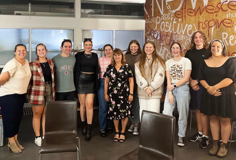 A group of 12 people huddled together and smiling in the corner of a room