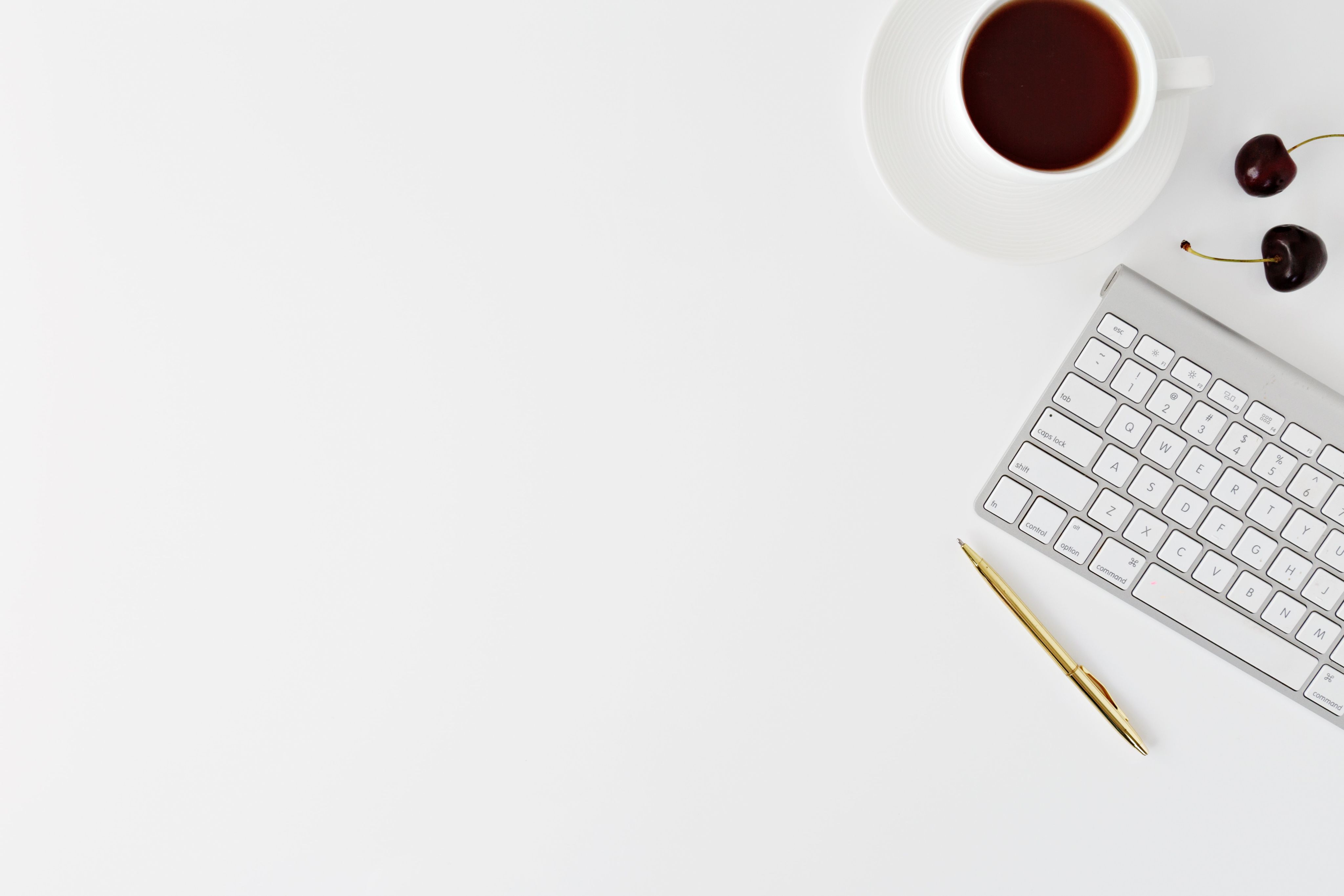 A white computer keyboard, a gold pen, a cup of black coffee and two cherries resting on a white desk