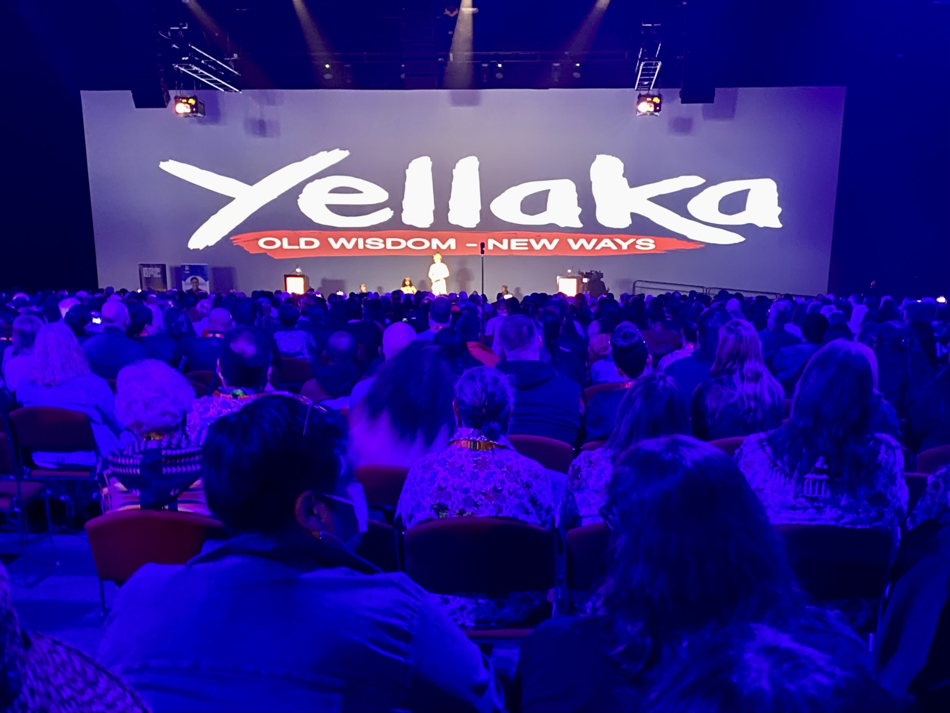 An audience at a conference. A large sign reading 'Yellaka: Old Wisdom, New Ways' is behind the presenters on the stage at the front of the audience.