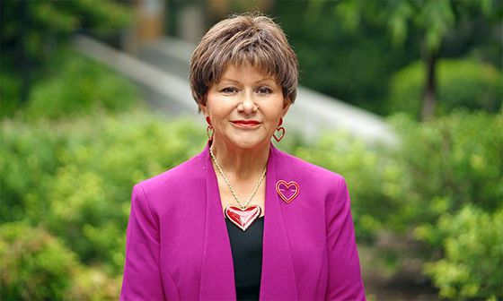 Professor Bronwyn Fredericks, a woman with short light brown hair, wearing a bright pink jacket and a large heart-shaped necklace. She is standing in front of green shrubs and is smiling slightly.