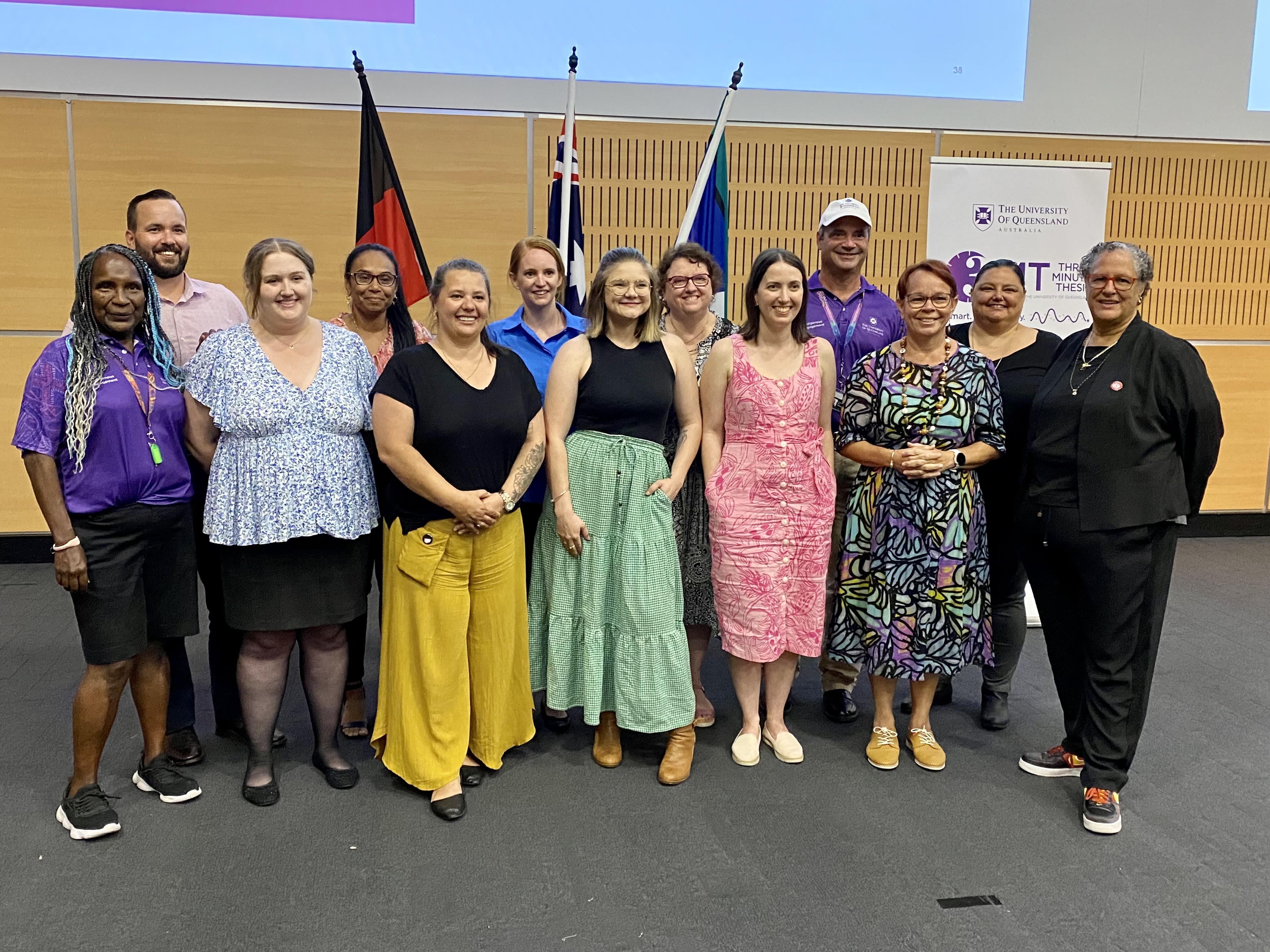 A group of 13 people standing in two rows in front of a light wood wall and the Aboriginal, Torres Strait Islander and Australian flags