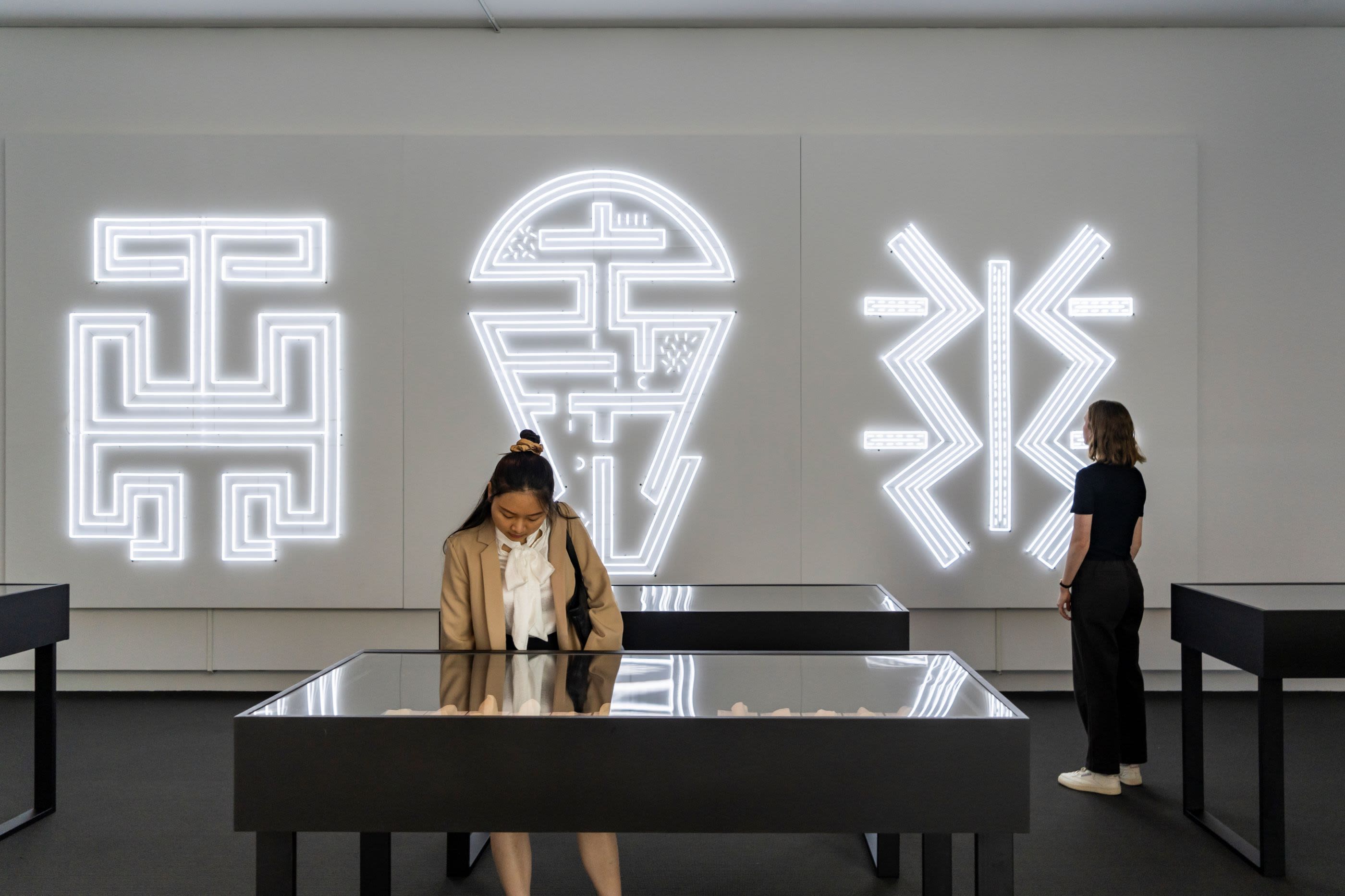 Two young women at an art exhibition with table display cabinets and symbols in lights on the wall behind them