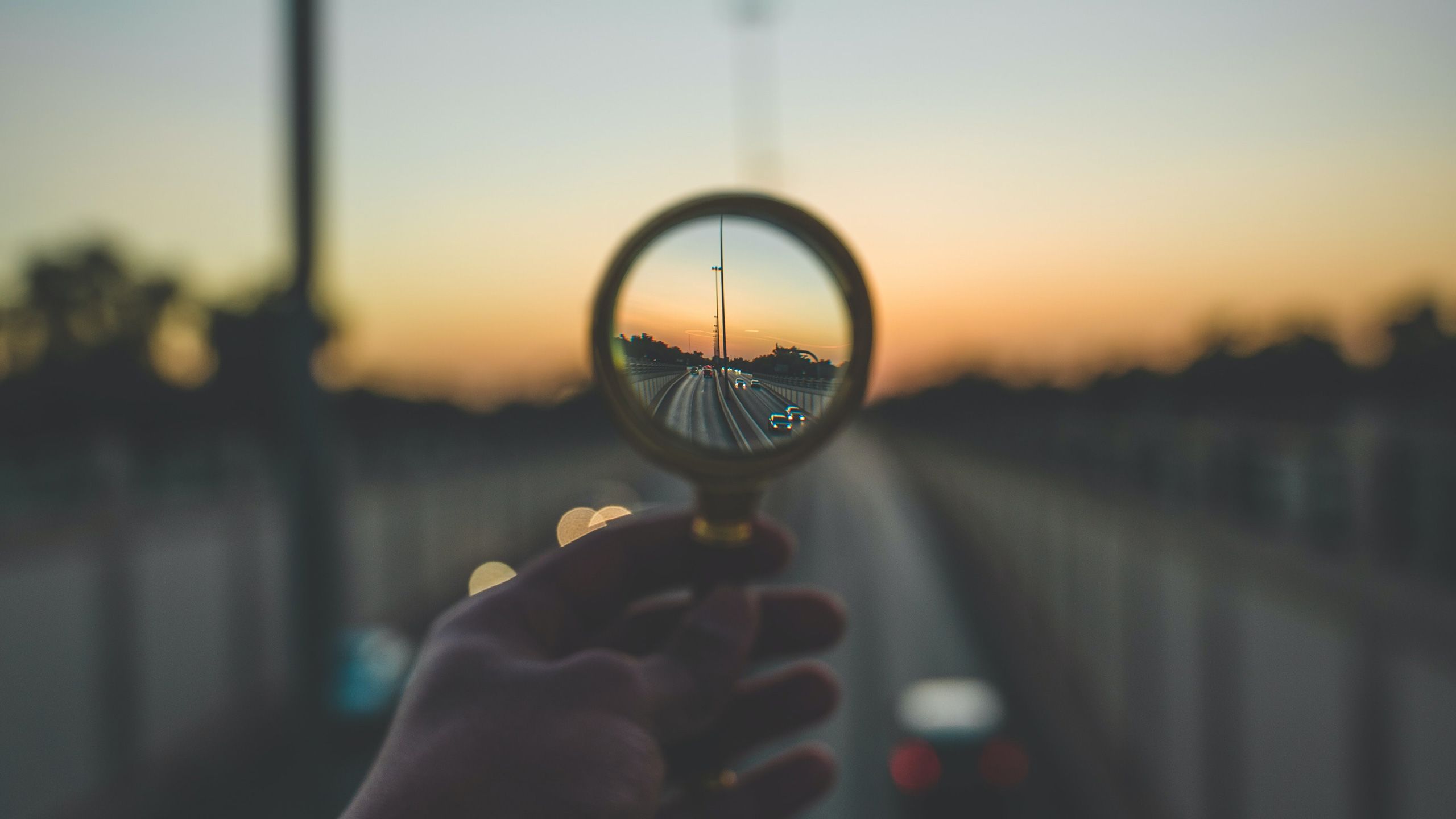 A hand holding a magnifying glass focusing on a sunrise over a road. The road is blurry except for the section being examined by the magnifying glass