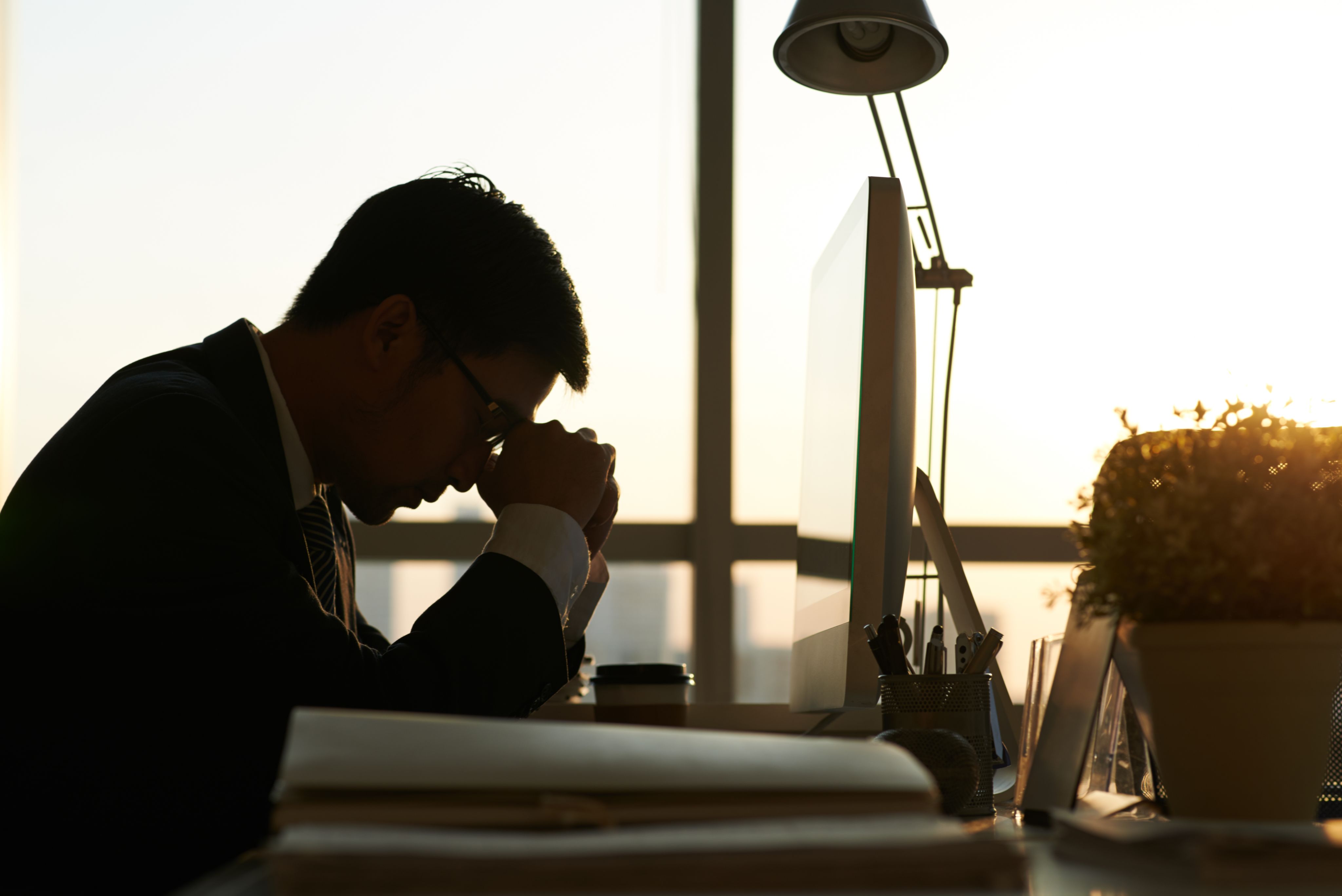Corporate worker seated at desk, leaning arms on it and resting his head against his hands.