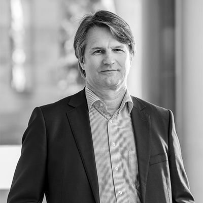 A black-and-white headshot of Dr Michael Collins, a middle aged man with light hair, smiling broadly while standing in front of a sandstone building