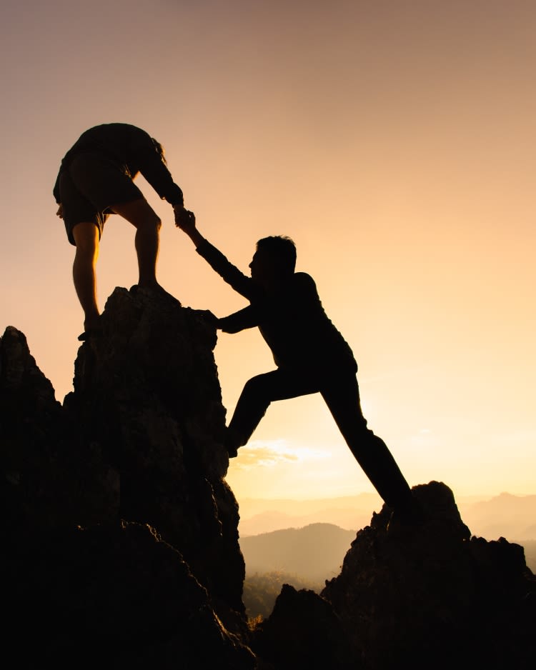 Silhouette of two male hikers climbing up mountain cliff and one of them giving a helping hand