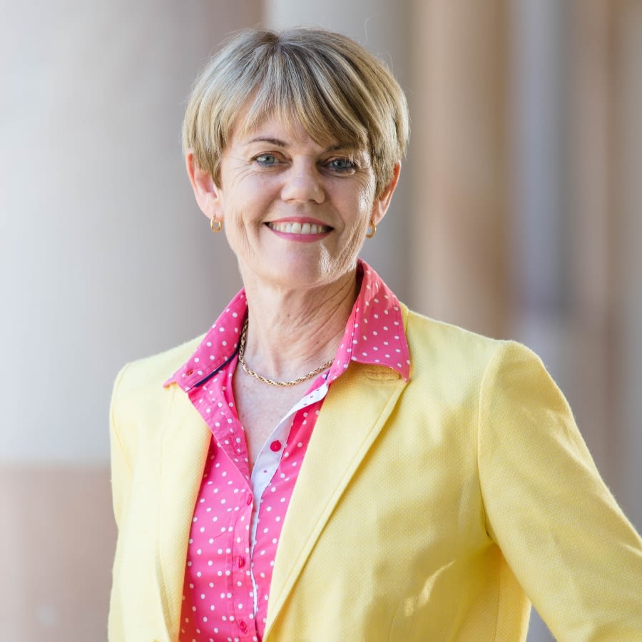 A headshot of Associate Professor Karen Hughes, a woman with short light hair, who is smiling while standing in front of a sandstone building