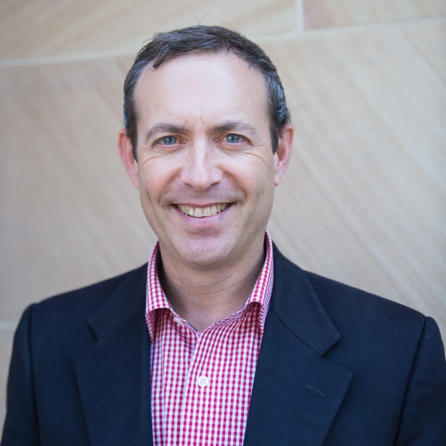 A headshot of Professor David Solnet, a middle aged man with short dark hair, smiling broadly while standing in front of a sandstone building