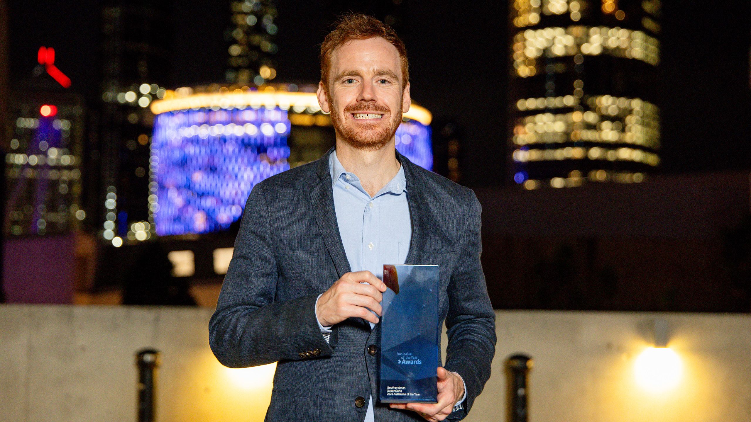 Geoff Smith standing with his Queensland Australian of the Year Award in front of bright city lights