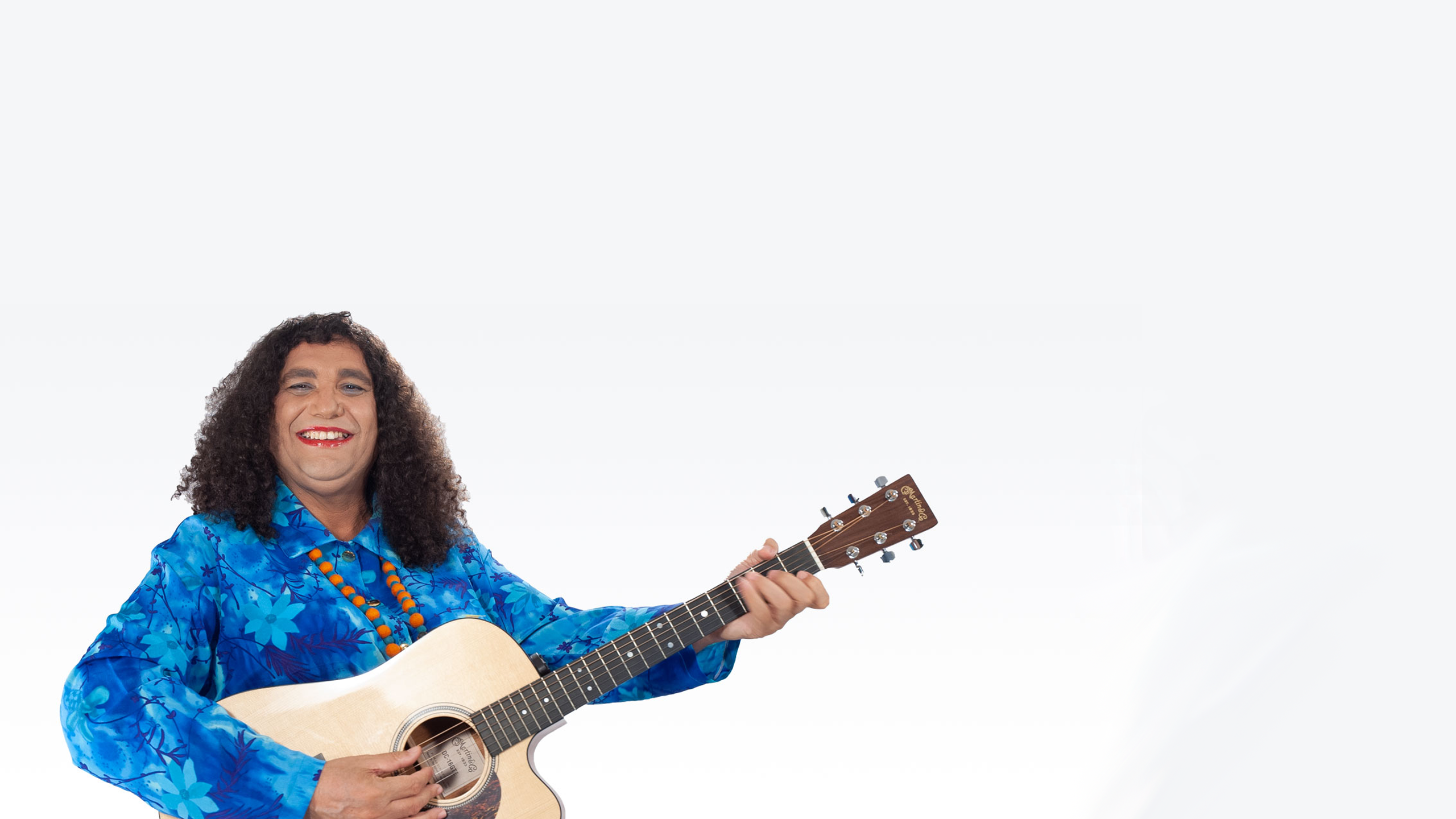 Image of a First Nations person with long curly hair, a bright blue long-sleeve shir and red lipstick - they are looking at the camera, smiling and holding an acoustic guitar.