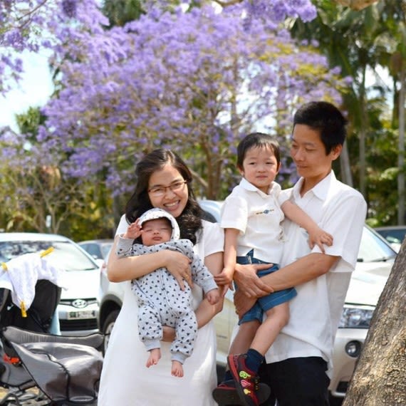 Phuong Tran standing next to her husband in front of UQ's jacaranda. She is holding her baby, and her husband is holding her other young son