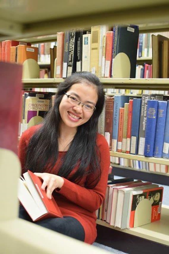 Phuong Tran holding a book while sitting in front of a shelf of books at the library