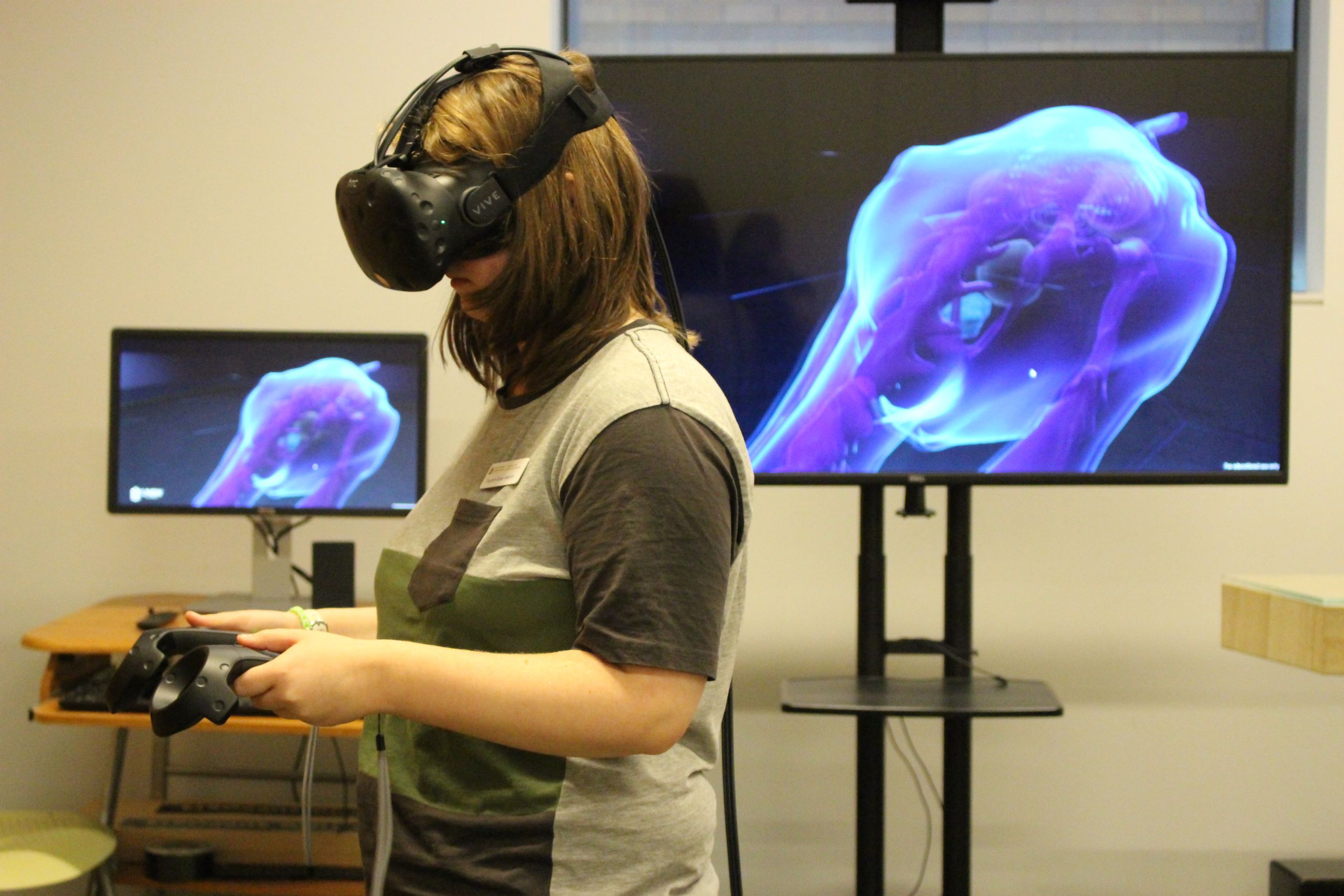 Female student operating VR controls while wearing the device over her eyes in front of a screen revealing what she is viewing.