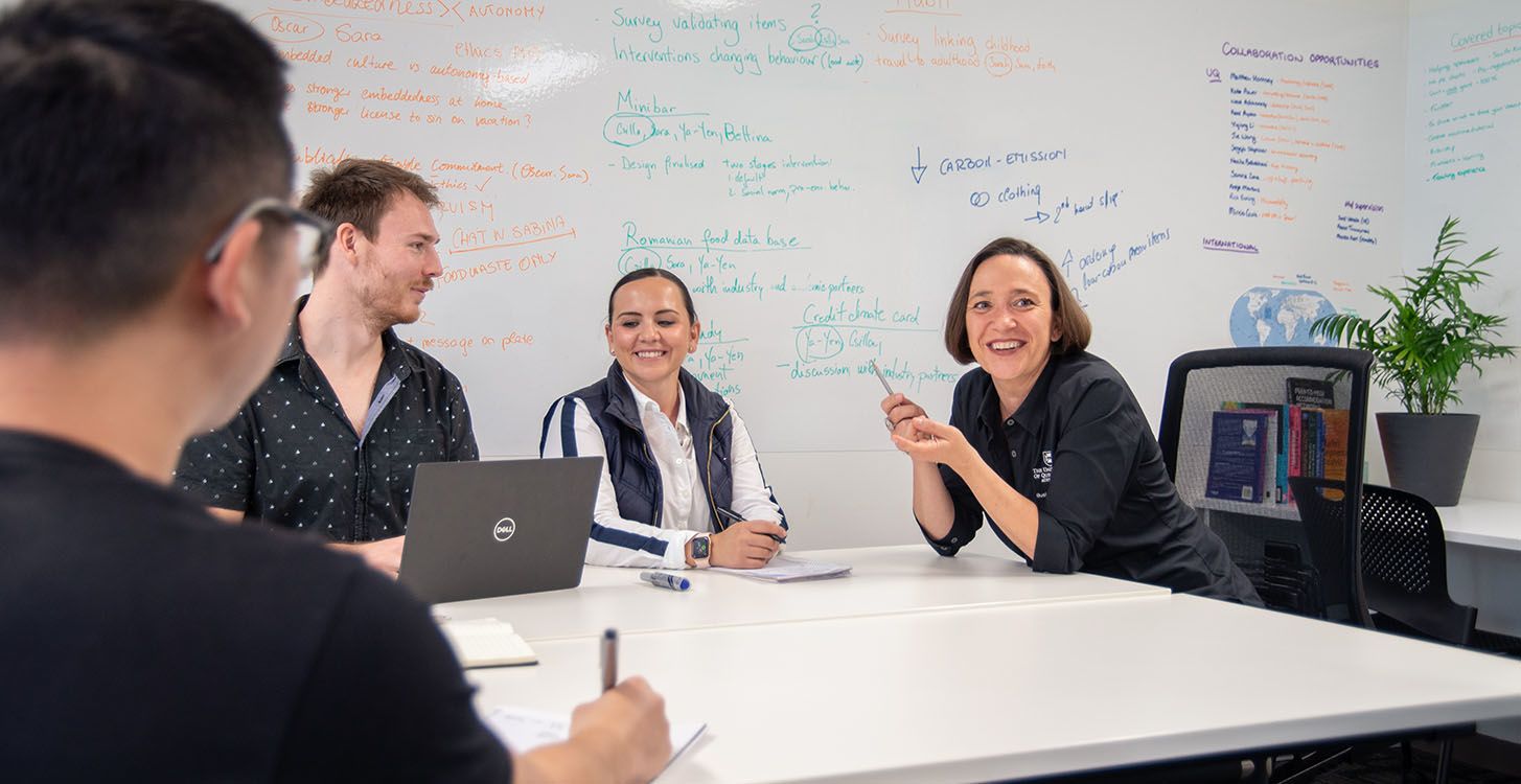 Sara Dolnicar sitting at a table with some of her proteges with a white board covered in writing behind them