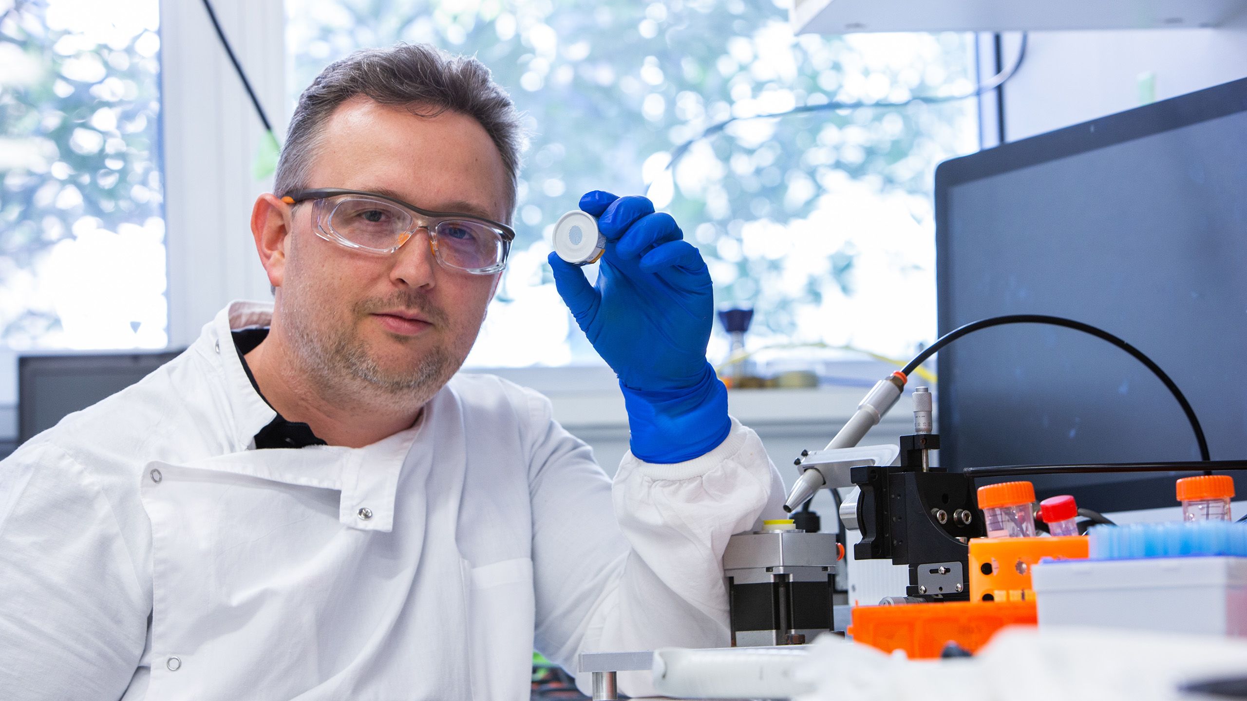 Dr David Muller wearing a white coat and blue gloves holding the high-density microarray patch up near his face as he faces the camera