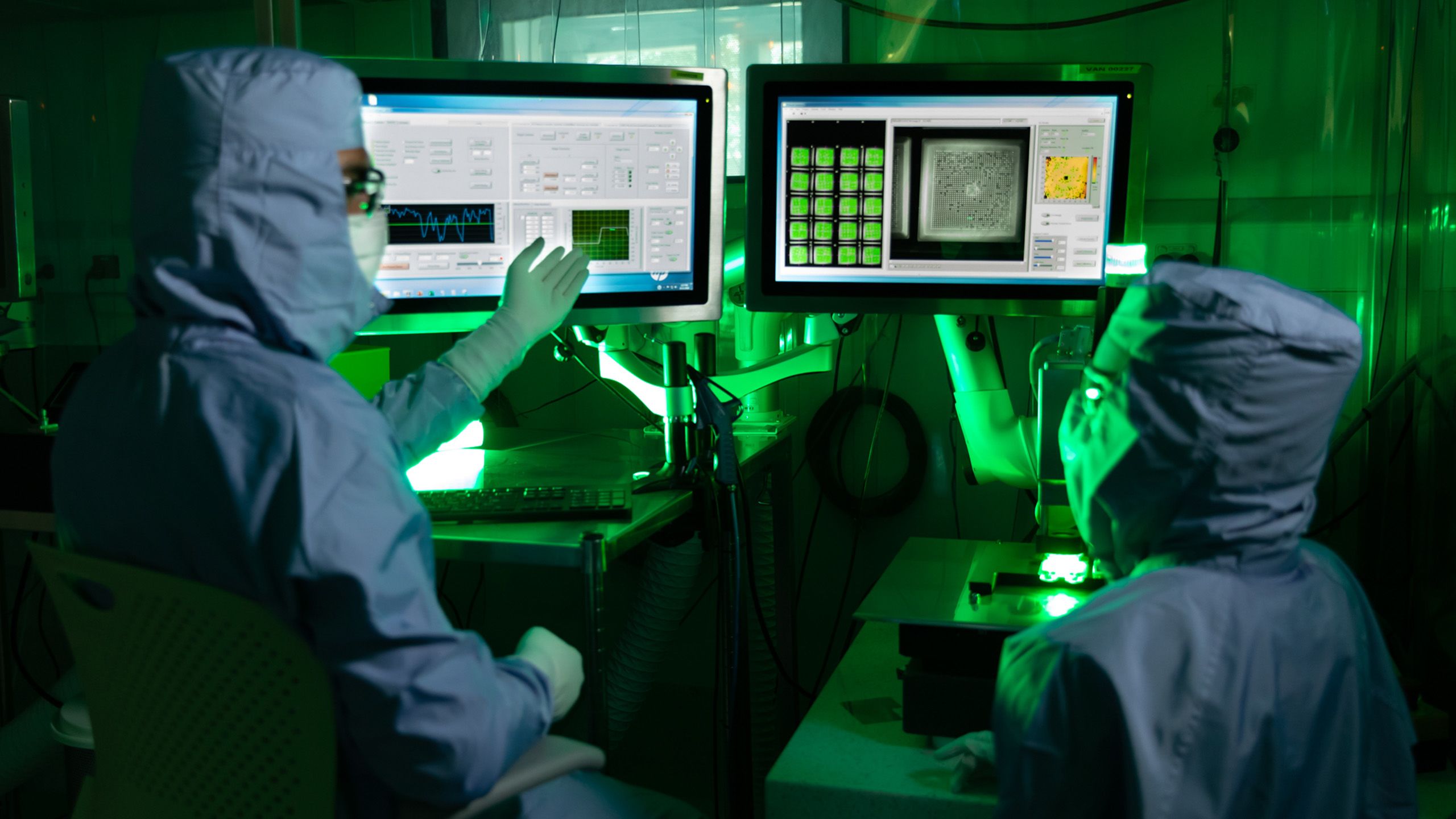 Researchers to left and right of screen wearing full protective gear, looking at a double computer screen, with green light backdrop 