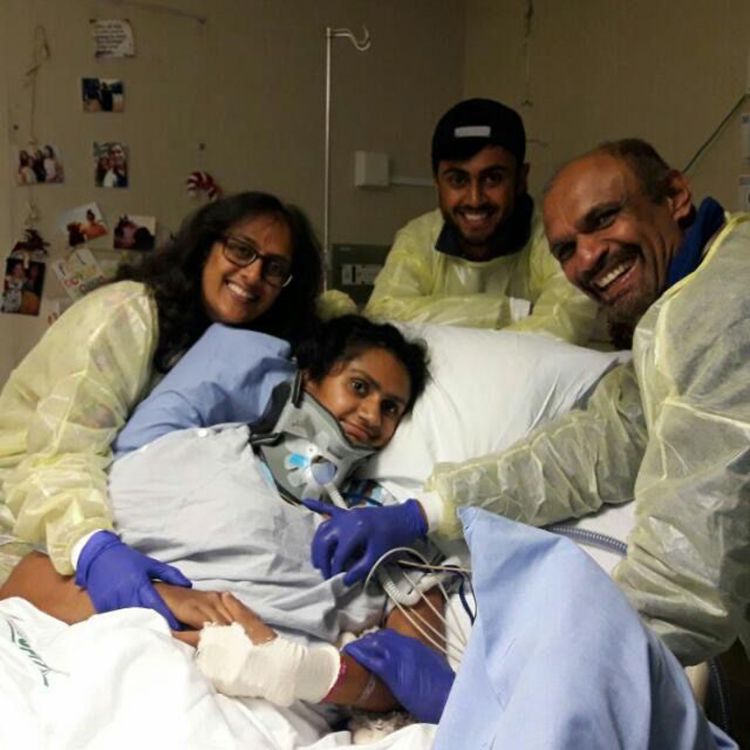 Family in protective clothing grouped around a patient in a bed wearing bandages and a neckbrace