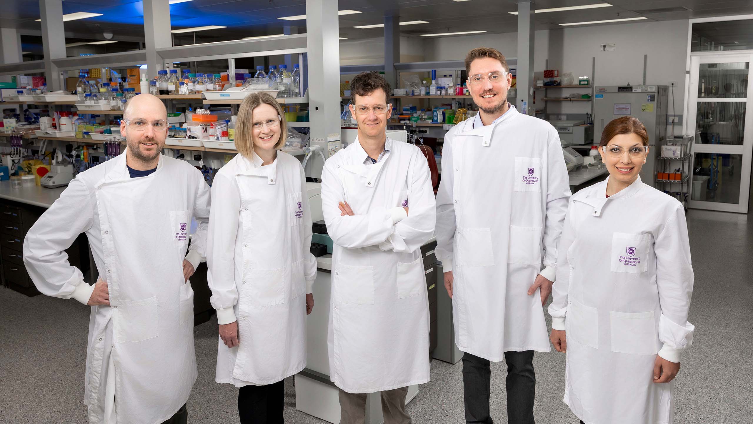Five scientists wearing UQ-branded lab coats in a laboratory. They are lined up in a row and smiling.