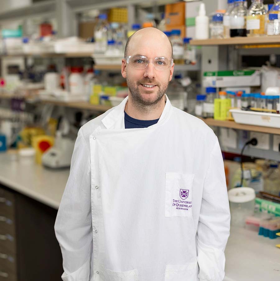 Associate Professor Daniel Watterson wearing a white coat and goggles standing in front of a row of desks and full shelves in a laboratory.